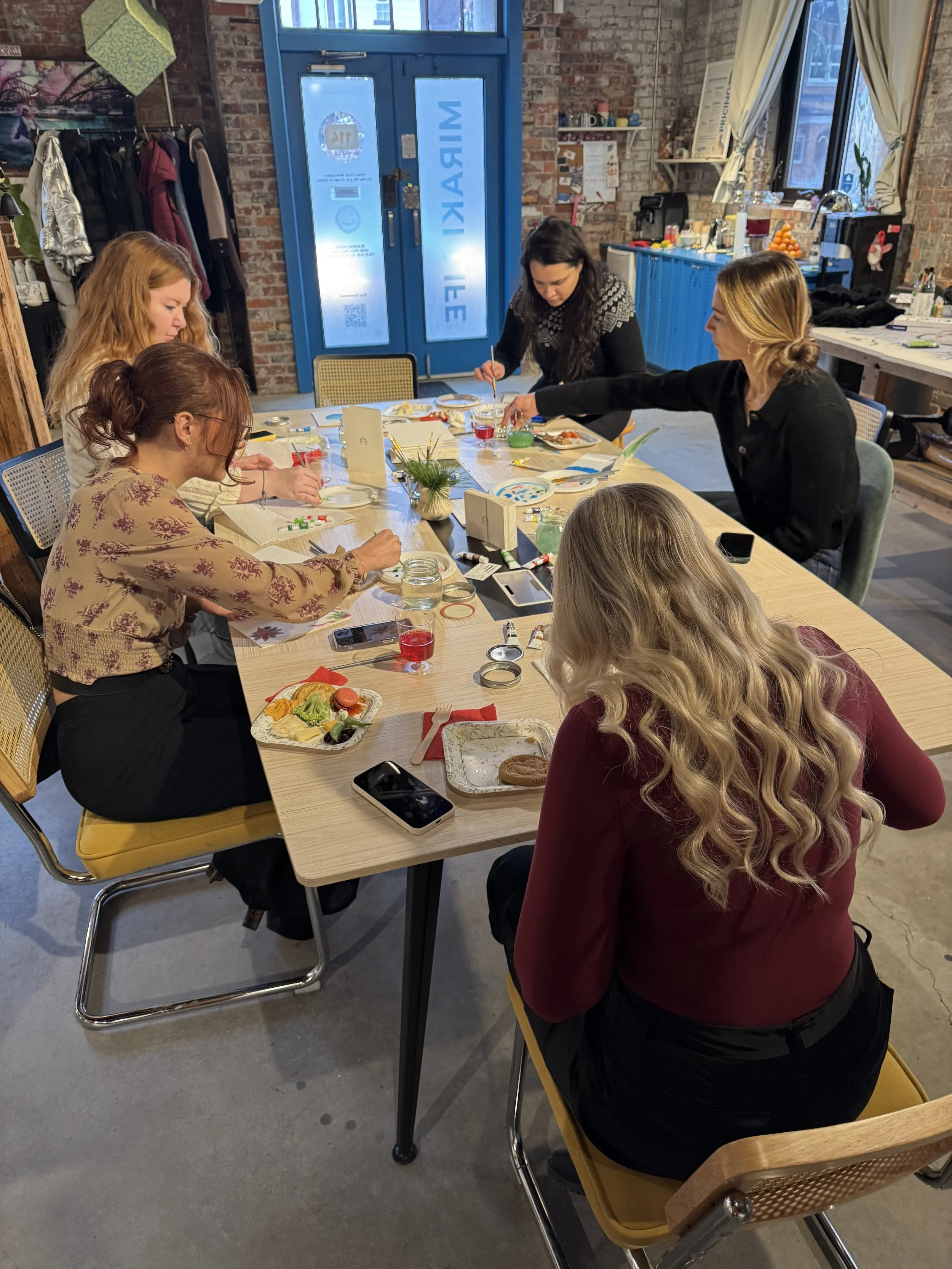 A group of six women sitting around a table engaged in arts and crafts activities with holiday-themed decorations in a cozy, industrial-style room that has exposed brick walls, large windows, and a table with snacks, drinks, and art supplies.