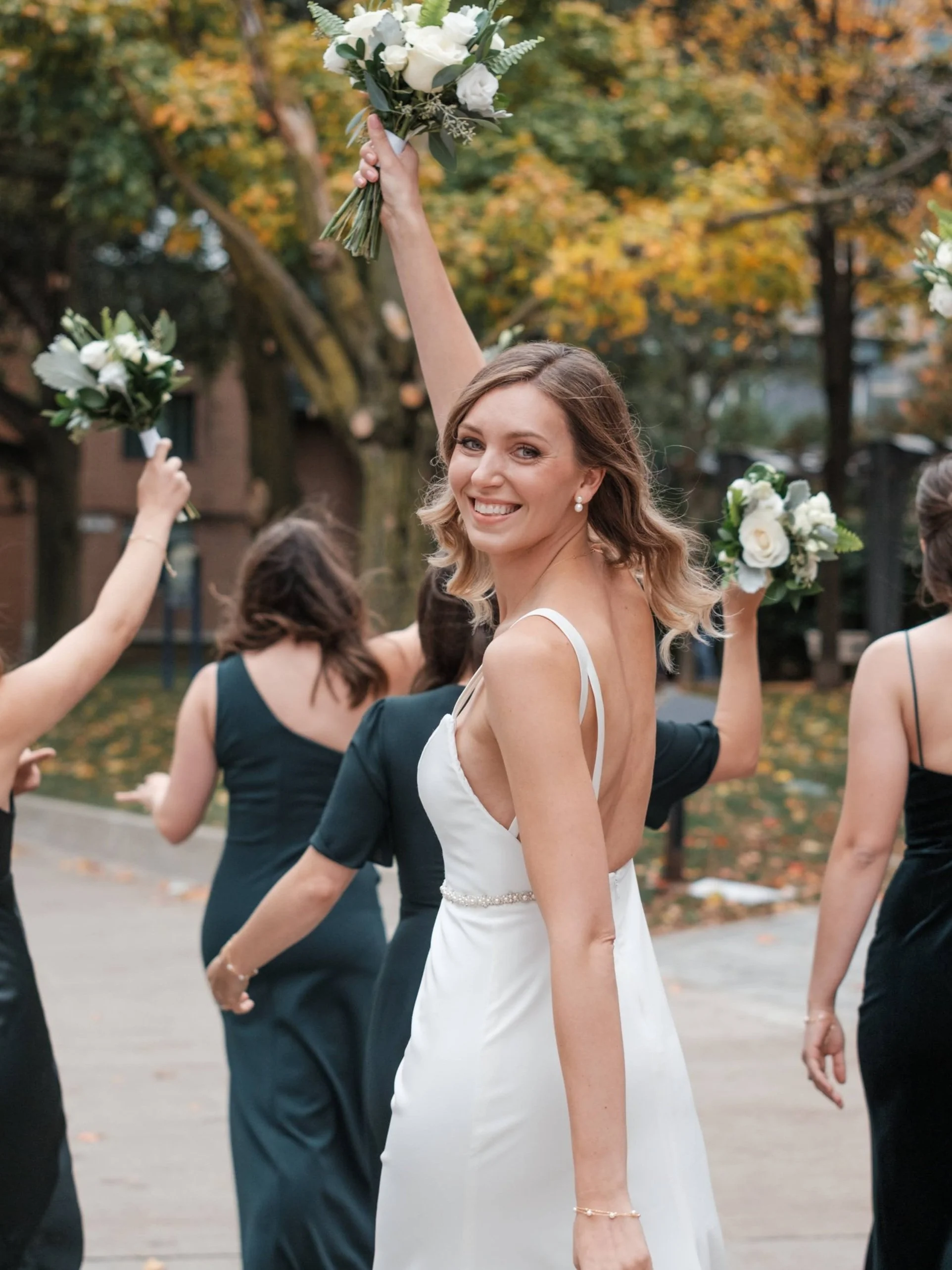 A smiling bride in a white wedding dress holding a bouquet in the air during a wedding celebration outdoors with autumn trees in the background.