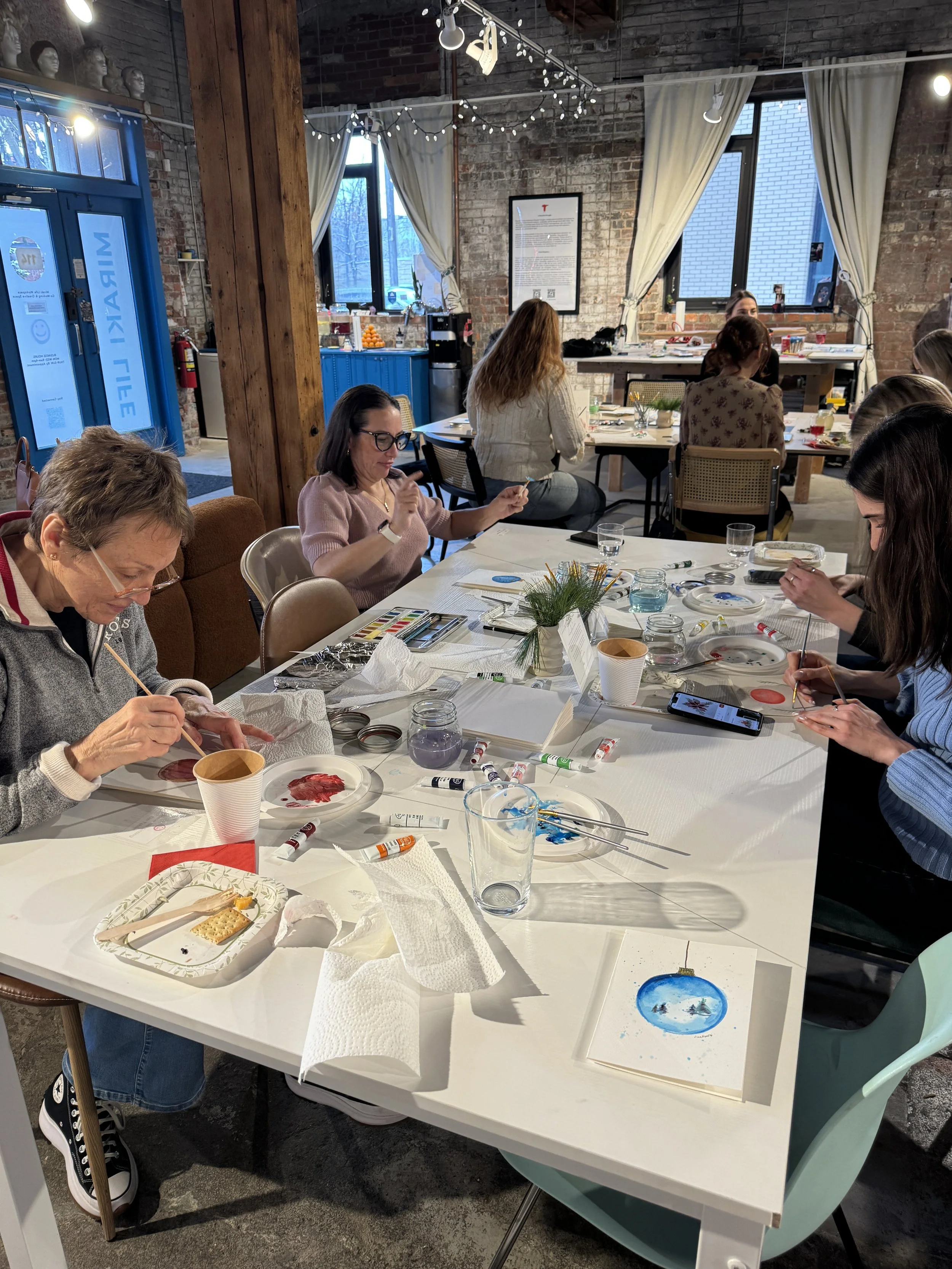Group of women participating in a paint and sip class at a decorated indoor venue with exposed brick walls, string lights, and large windows.