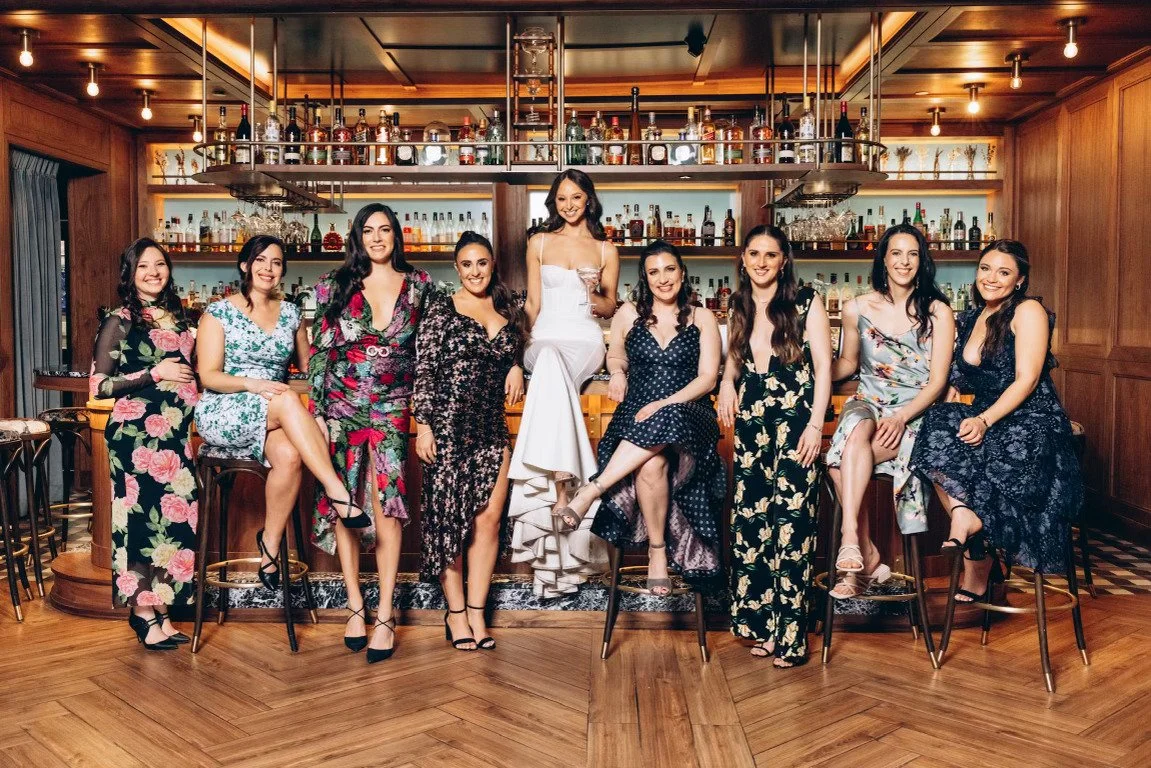 Nine women in dresses sitting on bar stools at a bar with liquor bottles behind them, smiling and posing for a group photo.