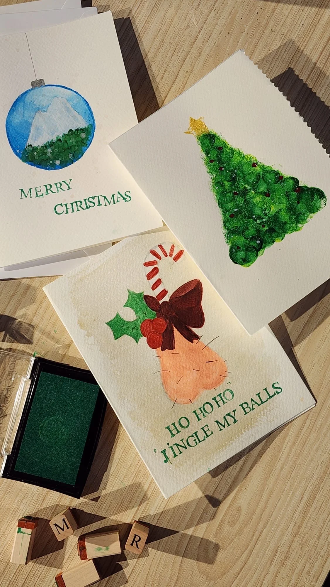 Three handmade Christmas cards featuring watercolor illustrations: a blue ornament with a mountain and green landscape, a Christmas tree with ornaments, and a Christmas carrot with holly and a bow. Small letter stamps and a green ink pad are also on the wooden surface.