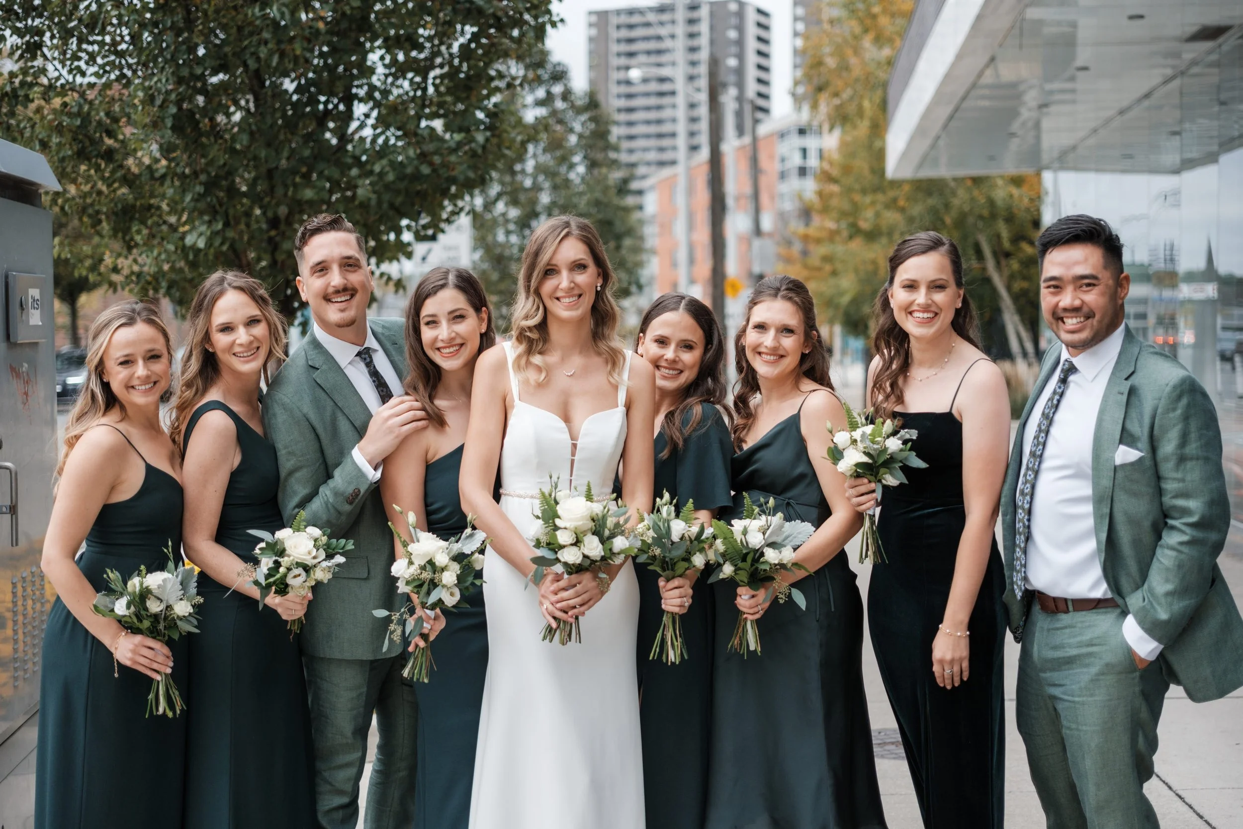 A group of nine people, including a bride in a white wedding dress holding a bouquet, and eight bridesmaids and groomsmen in formal attire, standing outdoors in an urban setting with tall buildings and trees in the background, smiling at the camera.