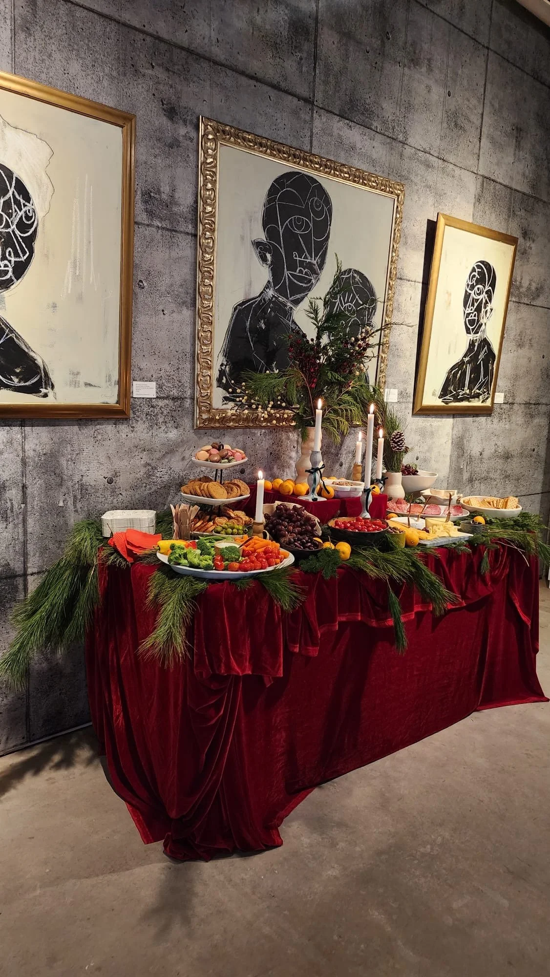 A decorated table with Christmas-themed treats and foods, adorned with pine branches and candles, in front of modern black-and-white art on a concrete wall.