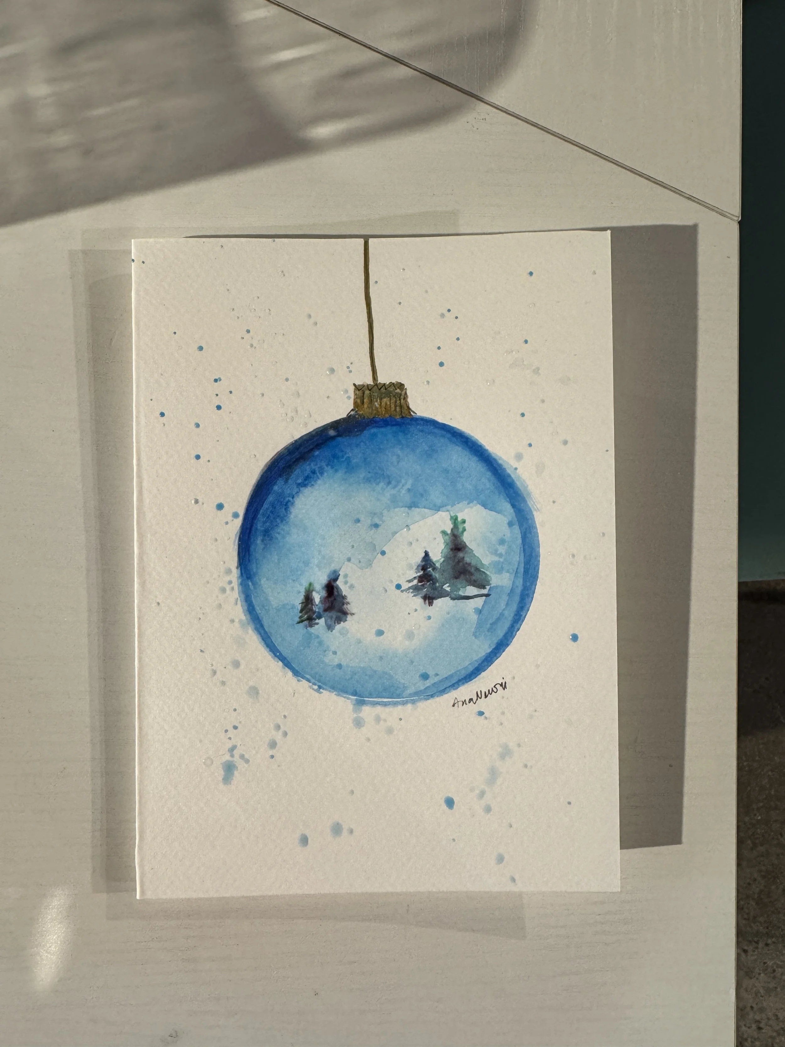 Watercolor painting of a blue Christmas ornament with a winter scene inside, featuring snow-covered trees. Signed by the artist Ananuragi.