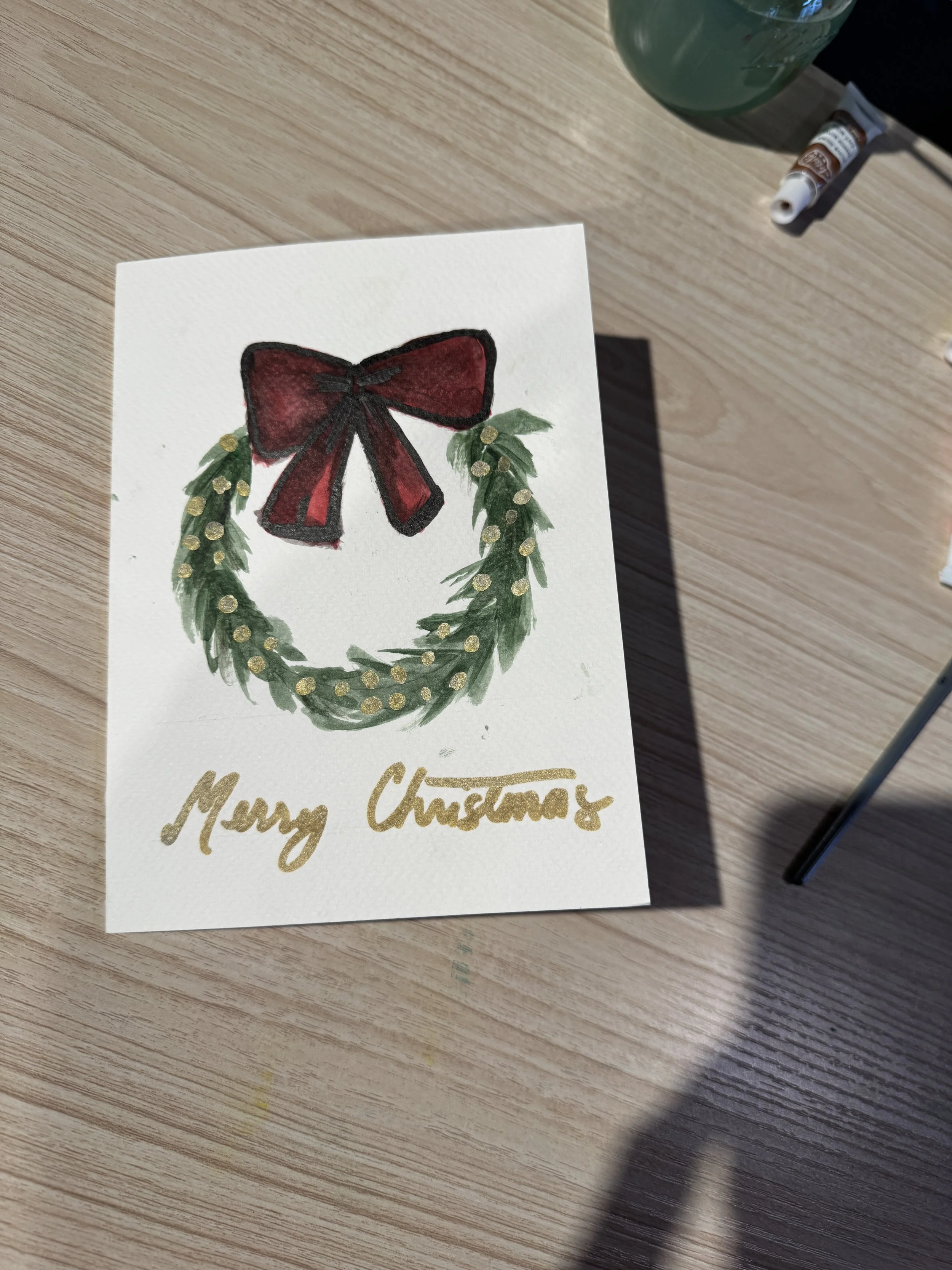 A handmade Christmas card with a green wreath decorated with gold dots and a large red bow on top. The words 'Merry Christmas' are written in gold at the bottom of the card. The card is placed on a wooden surface with some art supplies nearby.