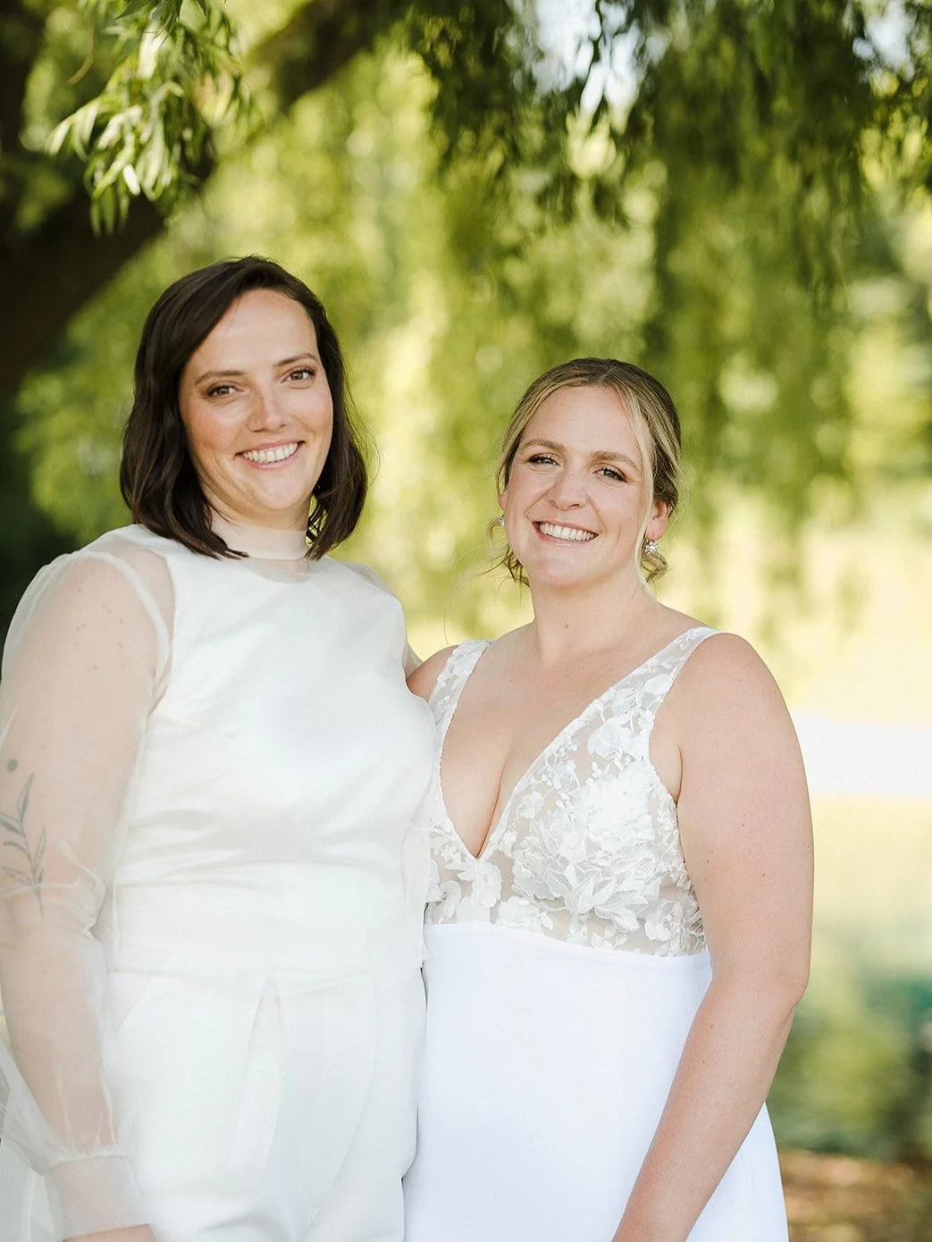 Two women in white dresses smiling outdoors with green trees in the background.
