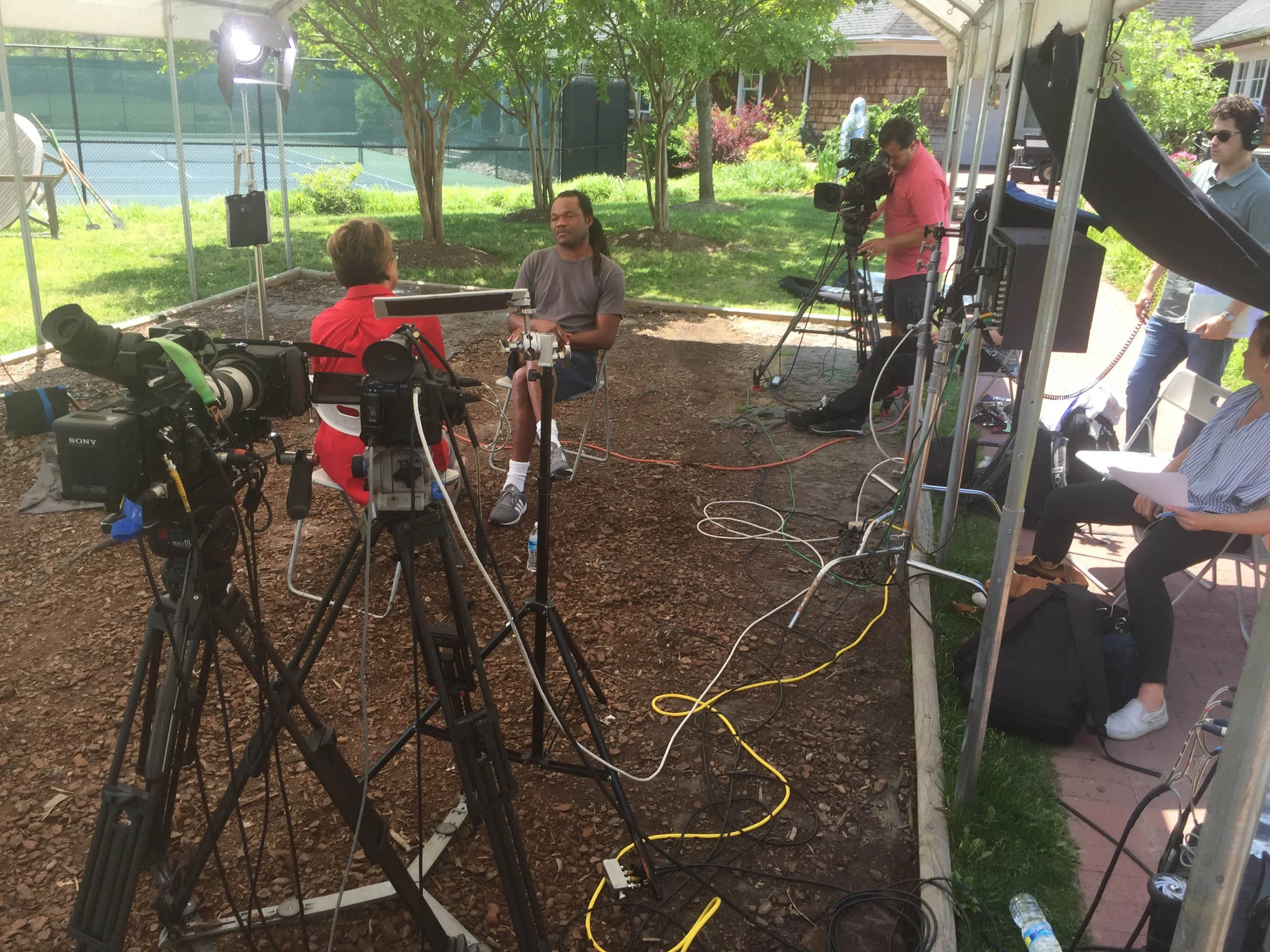 A backyard interview setup with two people seated, surrounded by cameras, lighting, and crew members filming in a shaded area with trees, a tennis court fence, and a house in the background.