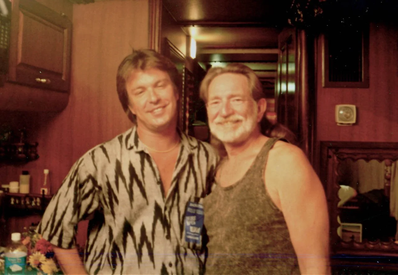 Two men standing close together in a warmly lit room, smiling at the camera. One man is wearing a patterned shirt, and the other is in a sleeveless top.