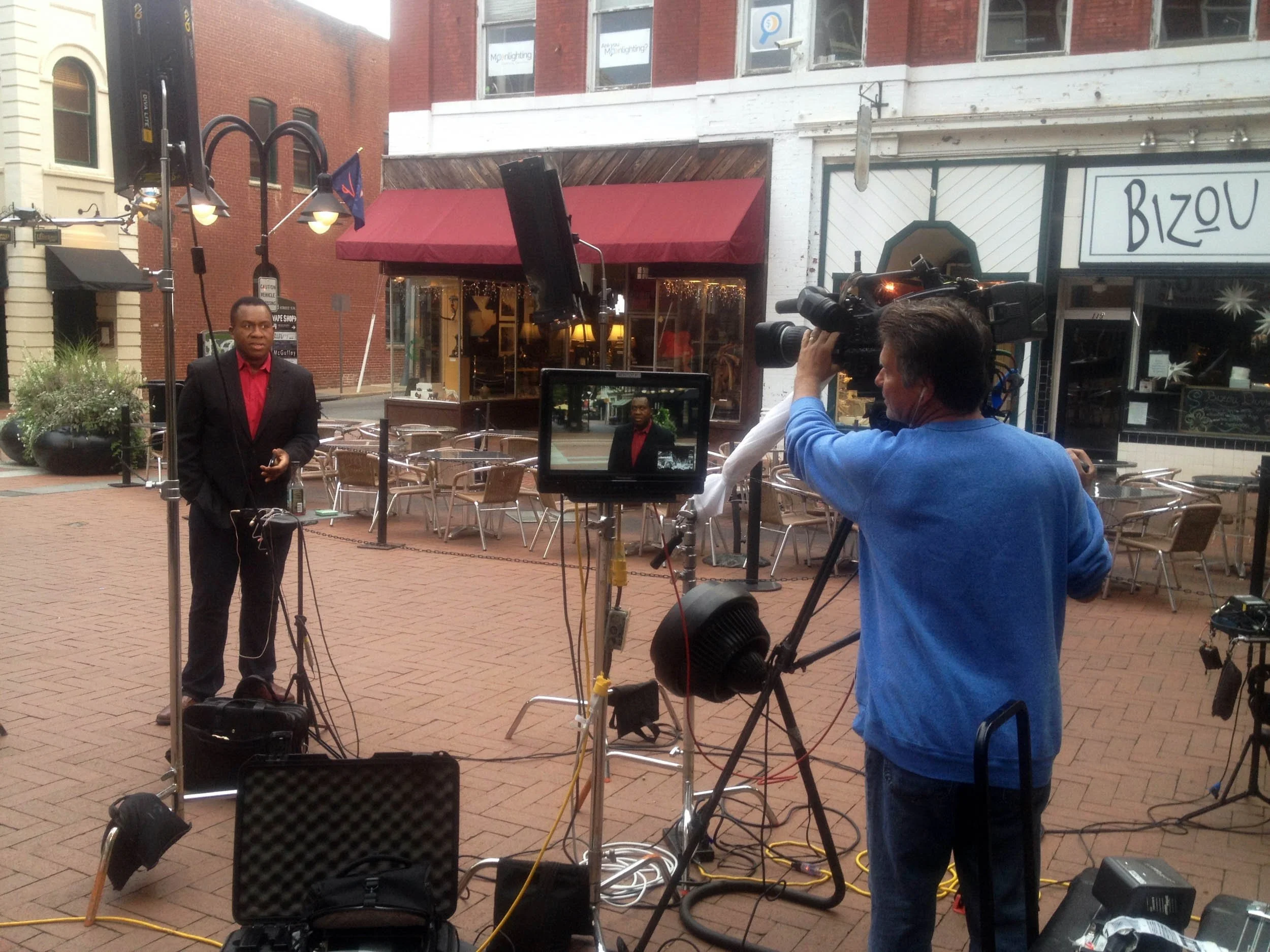 Television crew filming a man in a suit with a red shirt in an outdoor urban setting with empty tables and chairs.