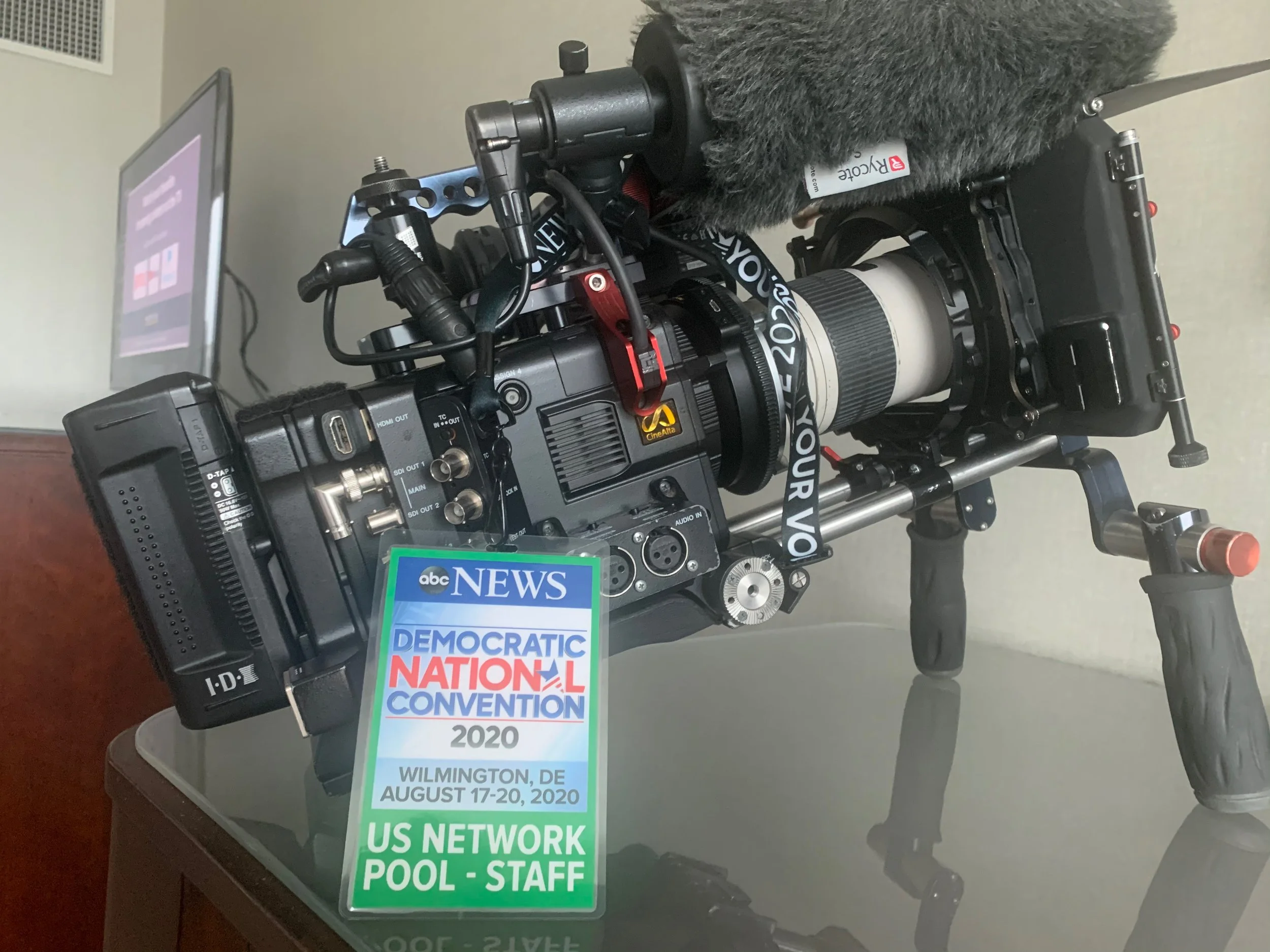 A professional video camera with a microphone and a monitor mounted on top, placed on a glass table, with a badge indicating coverage of the 2020 Democratic National Convention in Wilmington, Delaware.