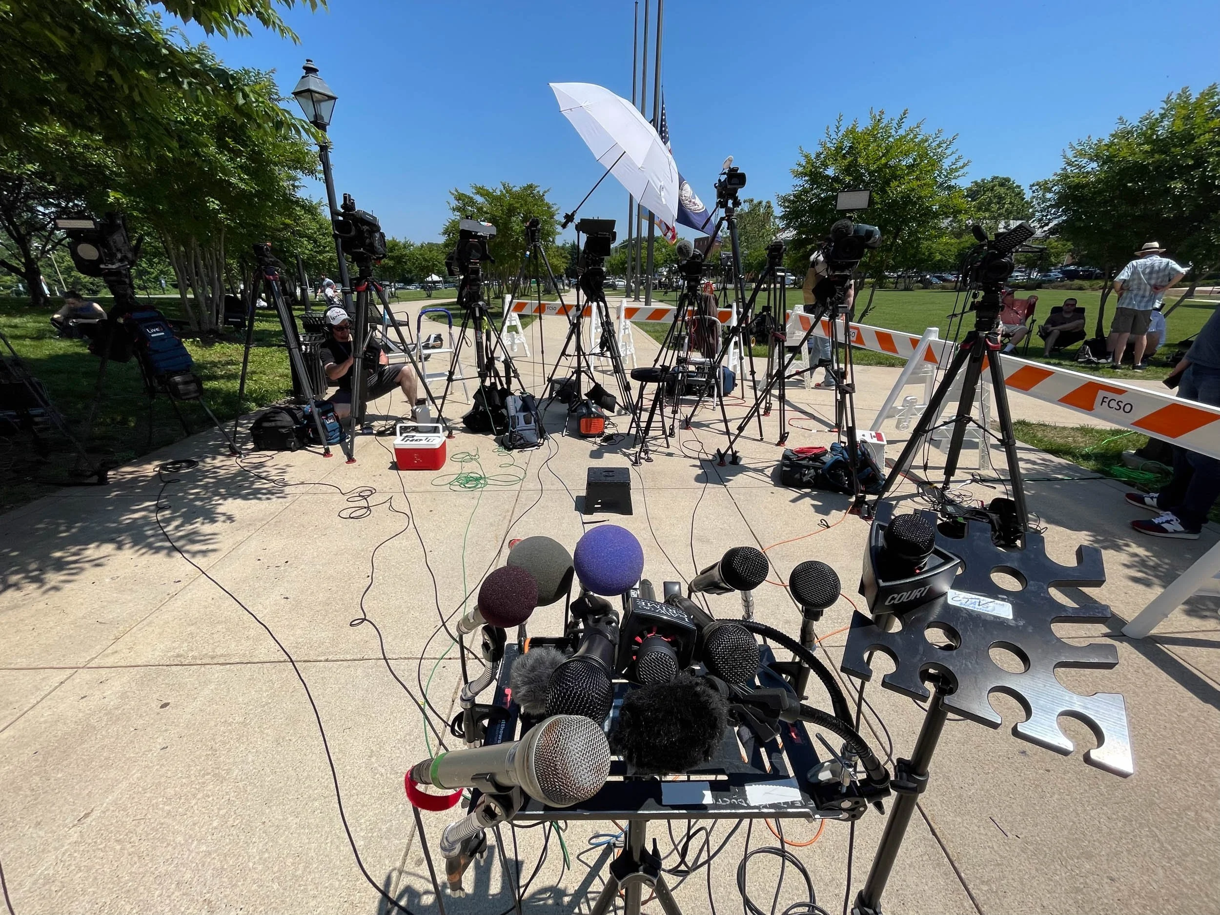A row of multiple cameras and tripods set up outdoors, possibly for a press or media event, with microphones and audio equipment in the foreground, and spectators sitting on benches in the background under clear skies and trees.