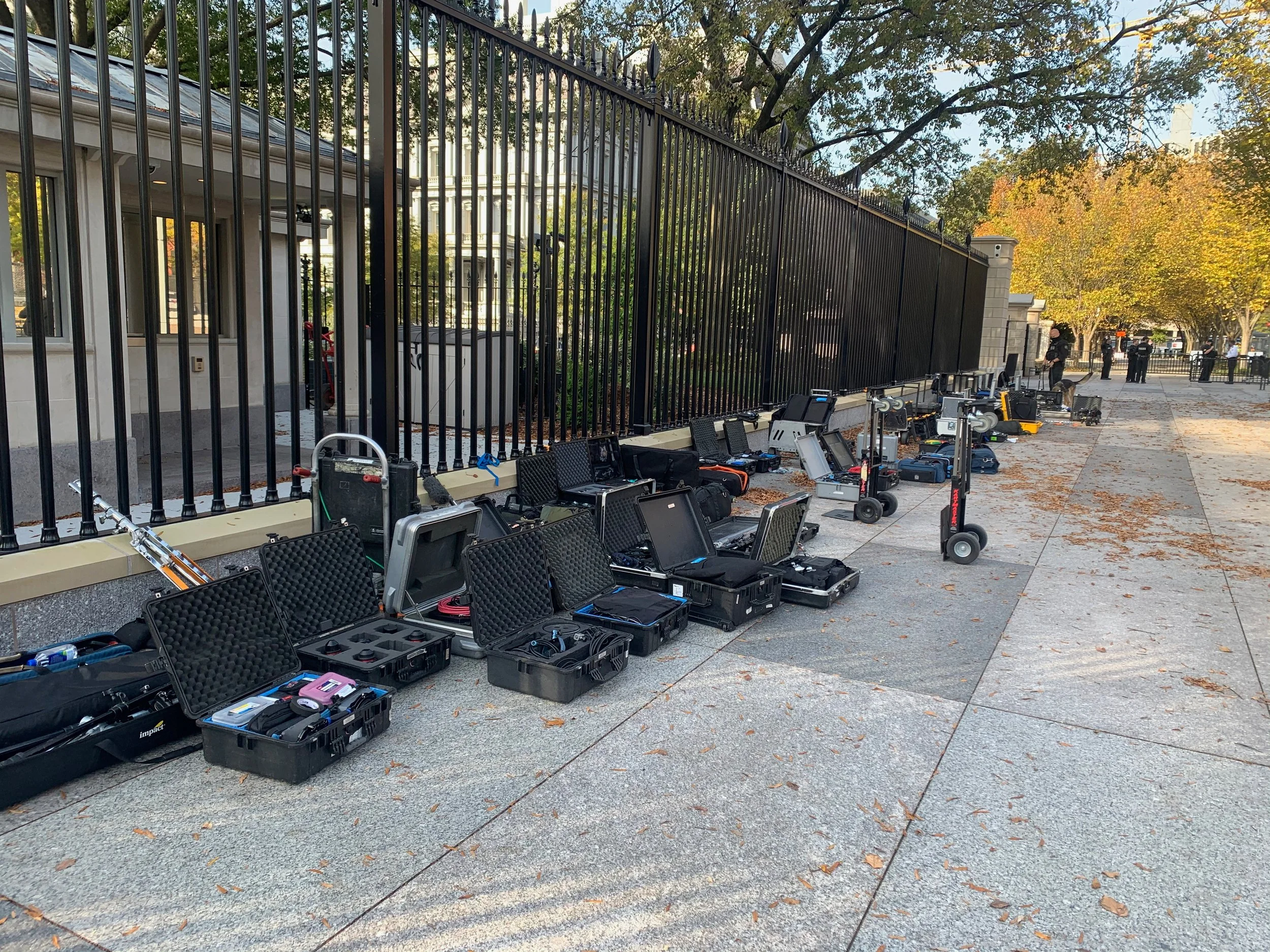 Several electronic equipment and tools are laid out in open cases on the sidewalk next to a black metal fence, with scooters among the equipment, and trees and some people in the background.