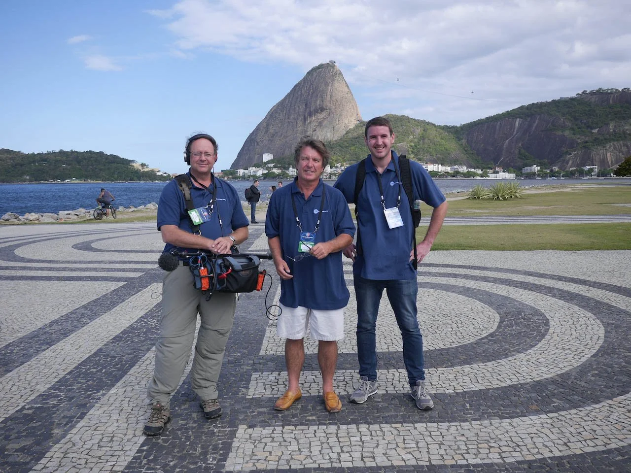 Three men standing outdoors on a patterned stone surface near a body of water, with Sugarloaf Mountain in the background, during daytime.