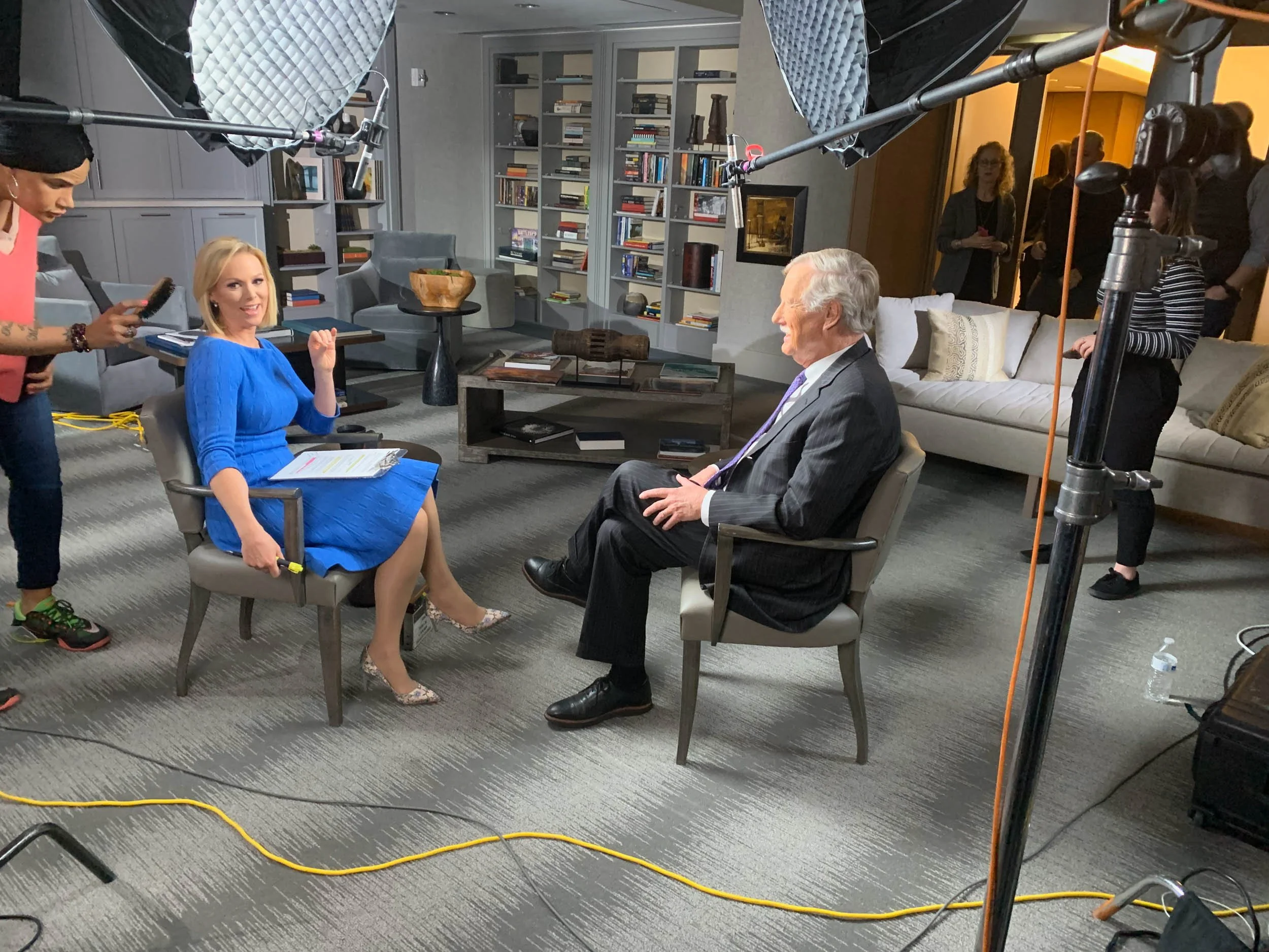 A woman in a bright blue dress being interviewed by a man in a suit in a professional interview setting with lighting equipment and crew members in the background.
