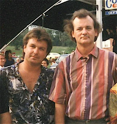 Two men standing outdoors at a fair or festival, with tents and other people in the background.