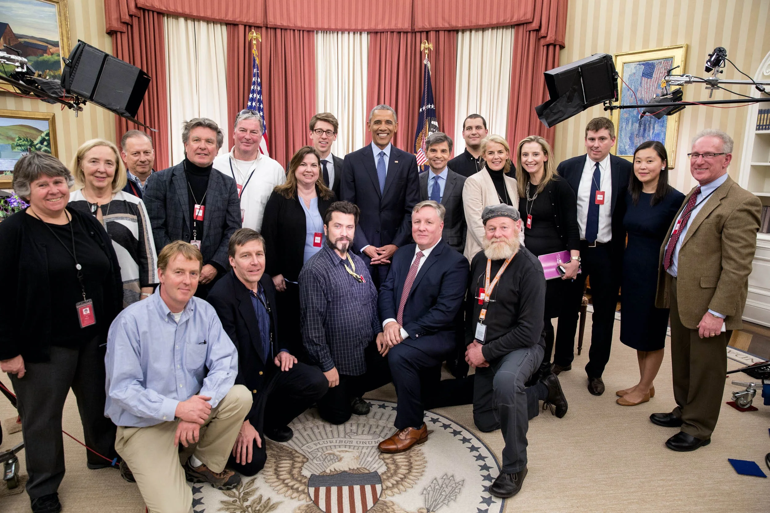 Group photo of diverse individuals, including former President Barack Obama, gathered indoors in front of the U.S. flag and presidential seal.