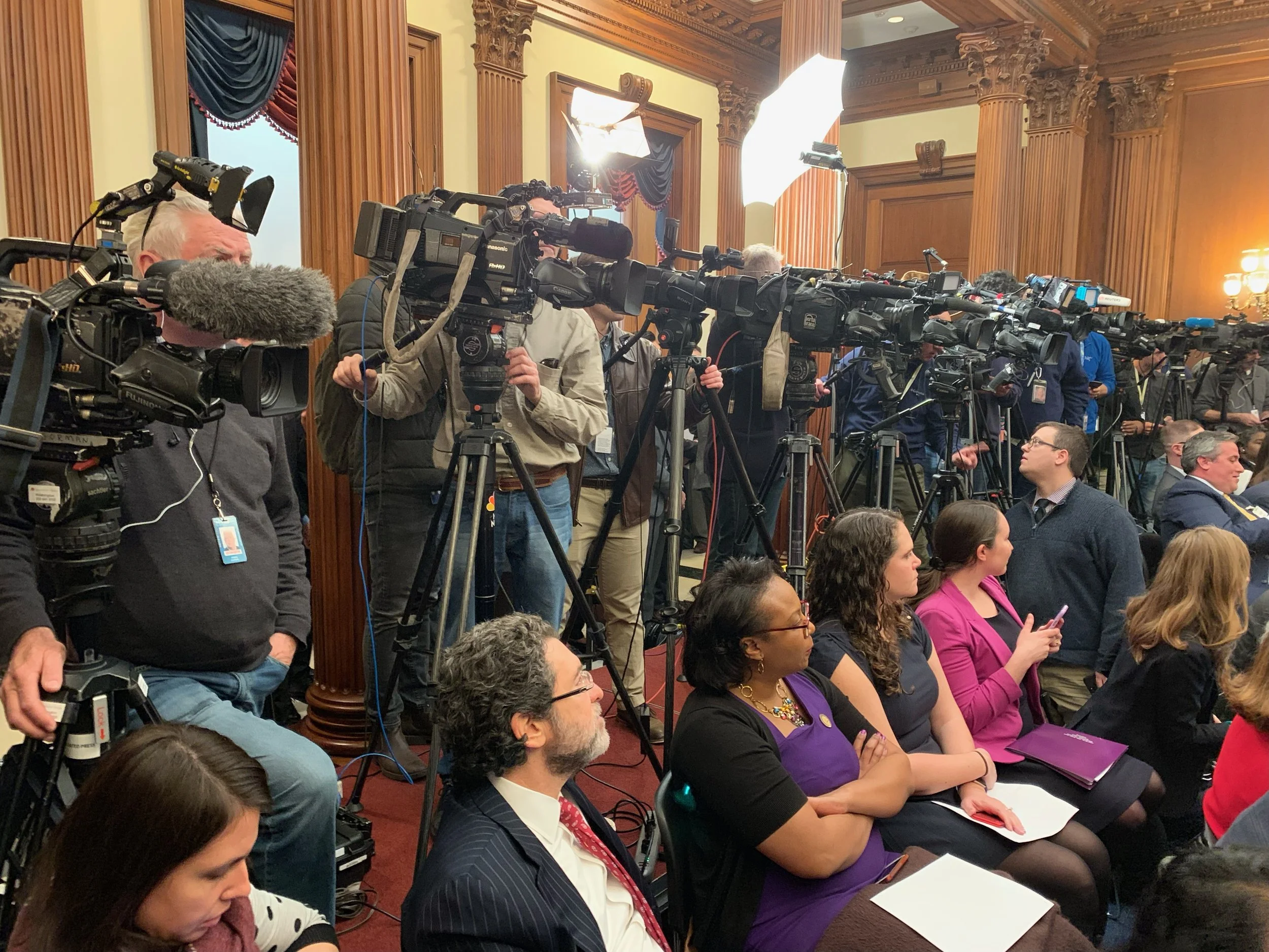 A room filled with numerous cameras and journalists during a press event or conference, with journalists sitting in the front row and large studio lights illuminating the scene.