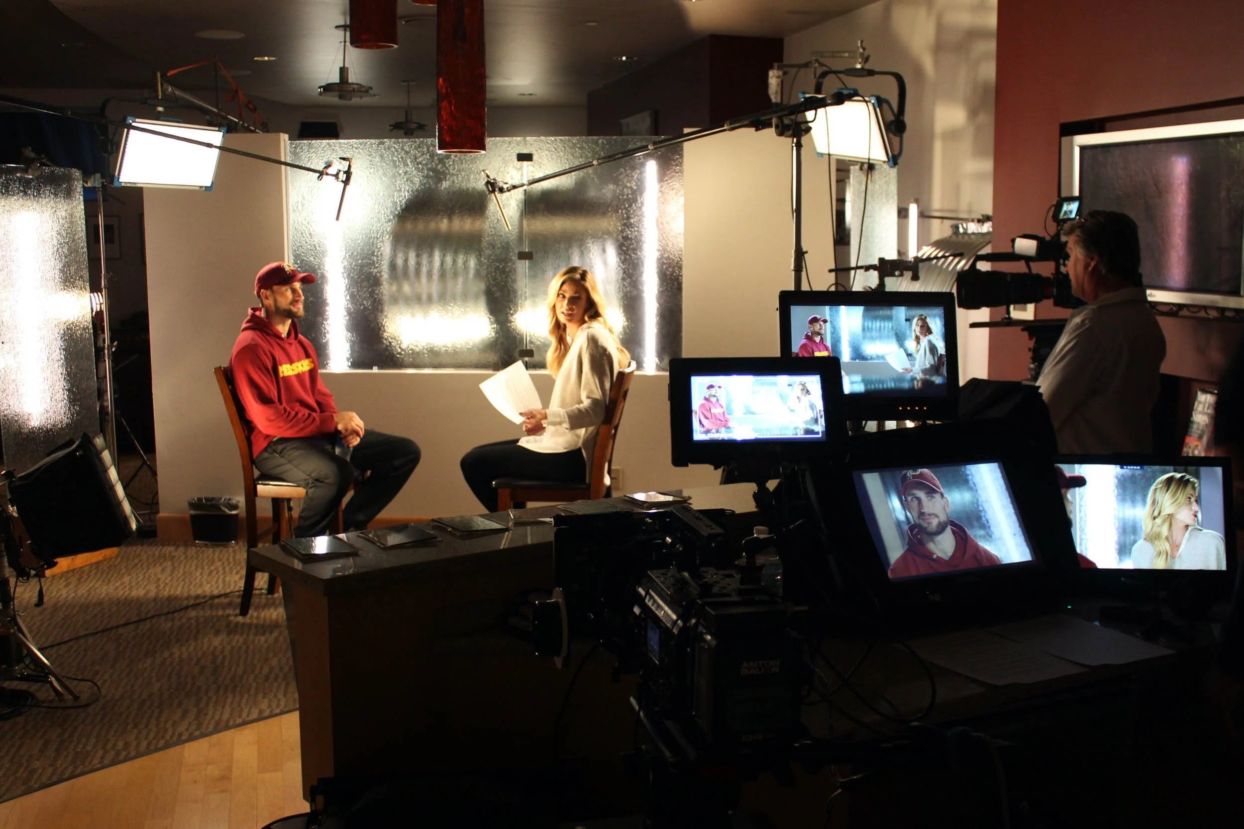 Behind the scenes of a video interview, with a man and woman talking on set, surrounded by cameras and lighting equipment, in a room with a reflective backdrop.