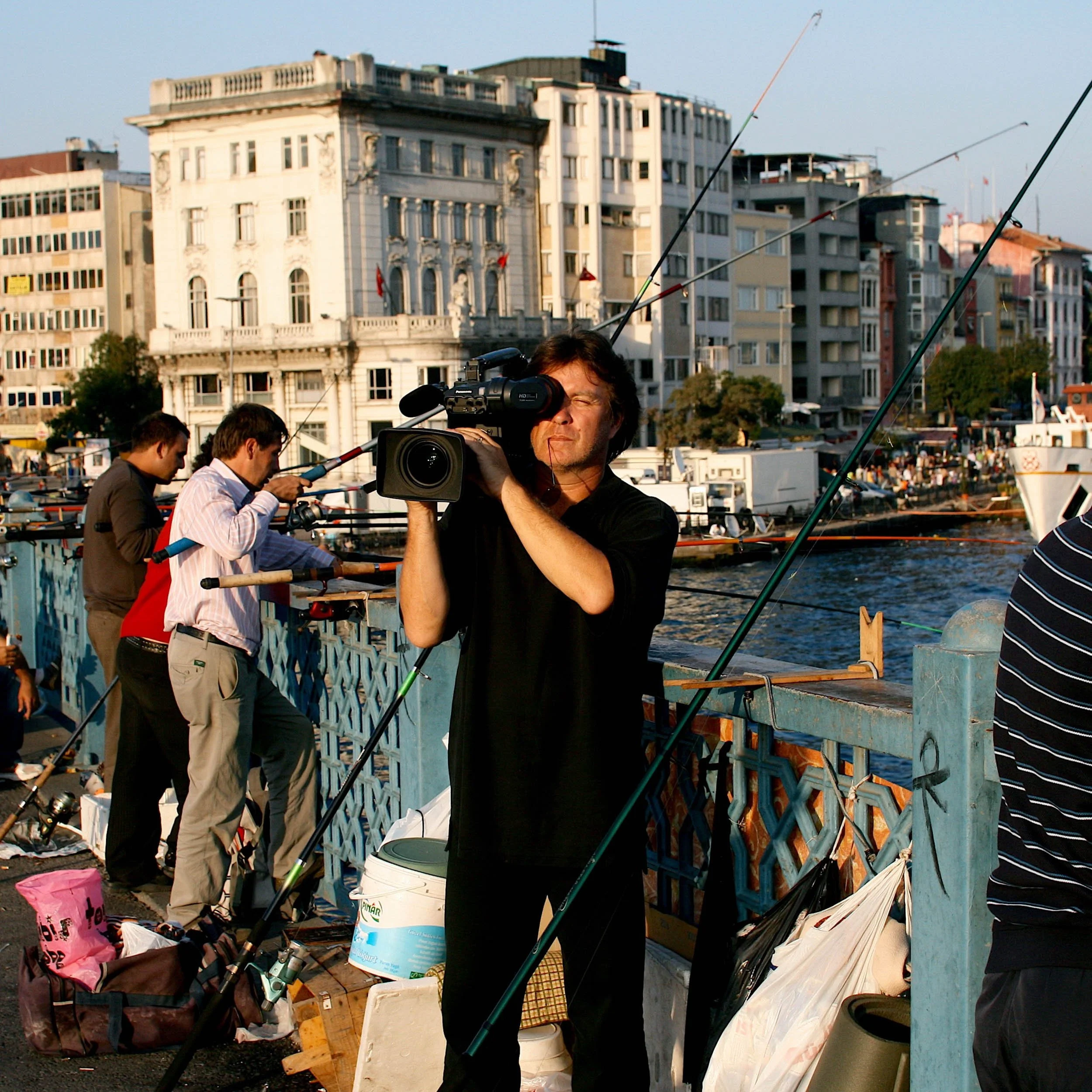 A man using a video camera to film near a waterfront with several fishing people and tall buildings in the background.