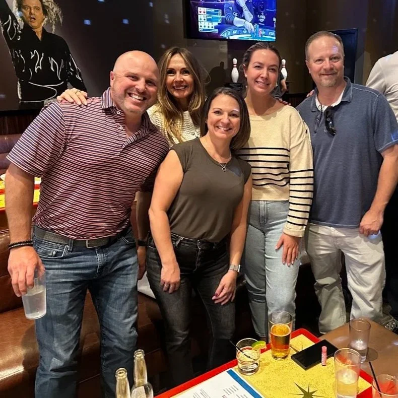 Group of seven people in a bar or lounge, smiling for the photo, some holding drinks, with a large screen and artwork in the background.