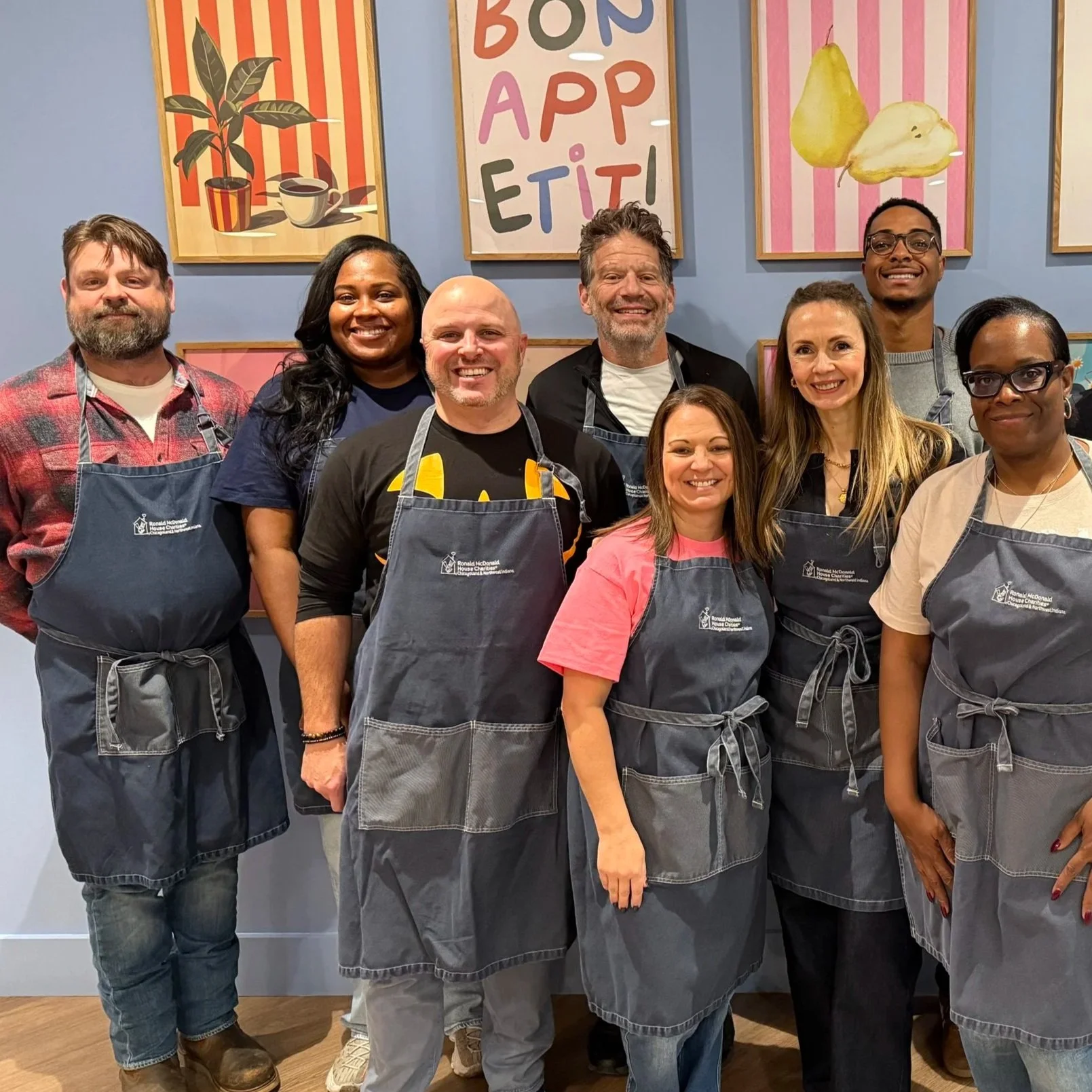 A group of ten diverse people wearing aprons posing indoors in front of colorful food-themed artwork on a light blue wall.