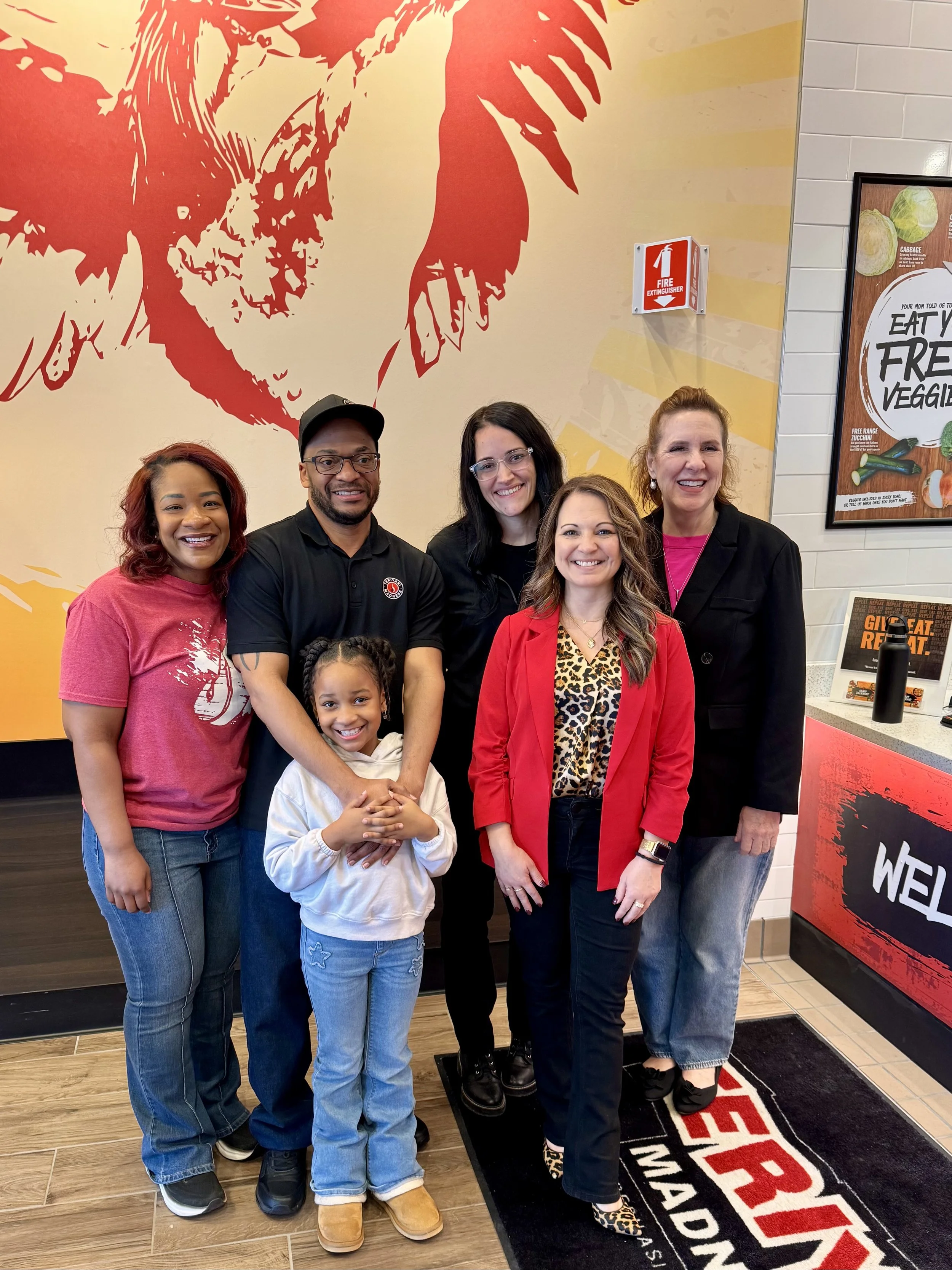 A group of six women and one young girl smiling and standing together inside a restaurant or fast-food place. The background features a large red and yellow graphic artwork of a chicken and some posters on the wall.