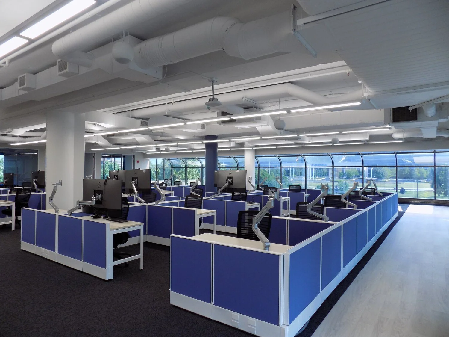 Empty modern office workspace with blue cubicles, black chairs, computer monitors, and flexible arms, large windows with city view, bright lighting, and industrial ceiling design.
