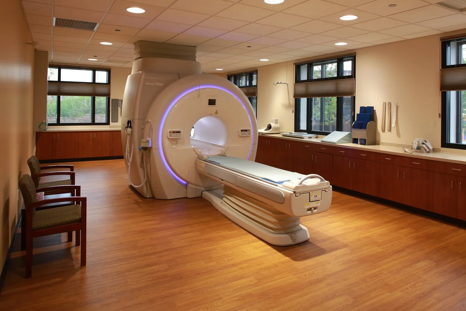 An empty hospital room with a large MRI machine in the center, wooden flooring, and multiple windows with blinds, along with chairs along the wall.