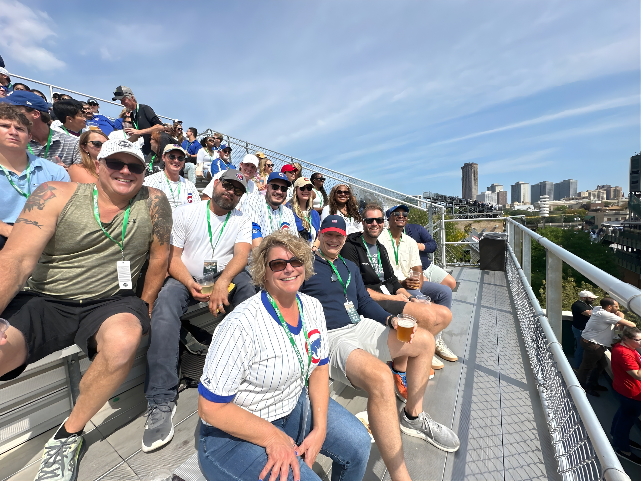 Group of people sitting on stadium bleachers, some holding drinks, smiling, with city skyline in the background on a clear, sunny day.