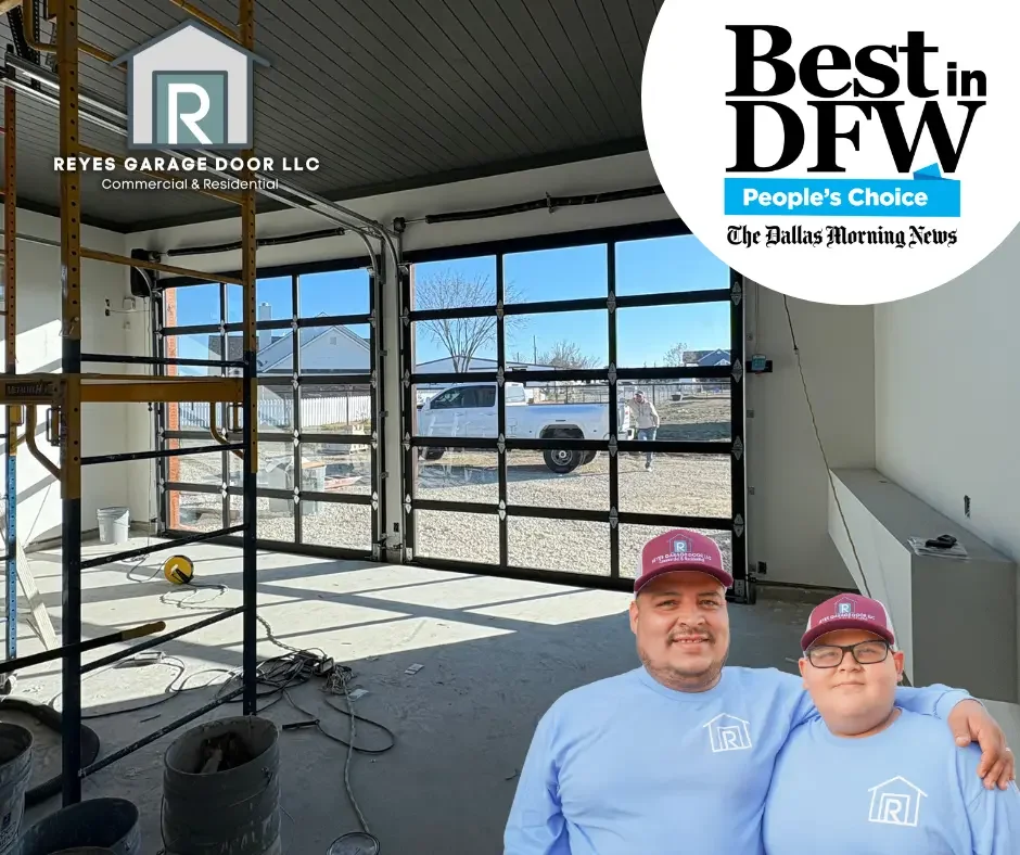Interior of a garage with a partially open black metal garage door, construction supplies, and two men wearing matching light blue shirts and maroon hats with a logo, standing inside and smiling.