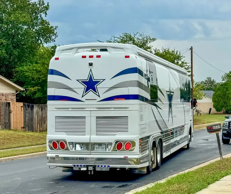 A white bus decorated with blue and gray star-themed graphics parked on a suburban street, with a mailbox and other vehicles visible.