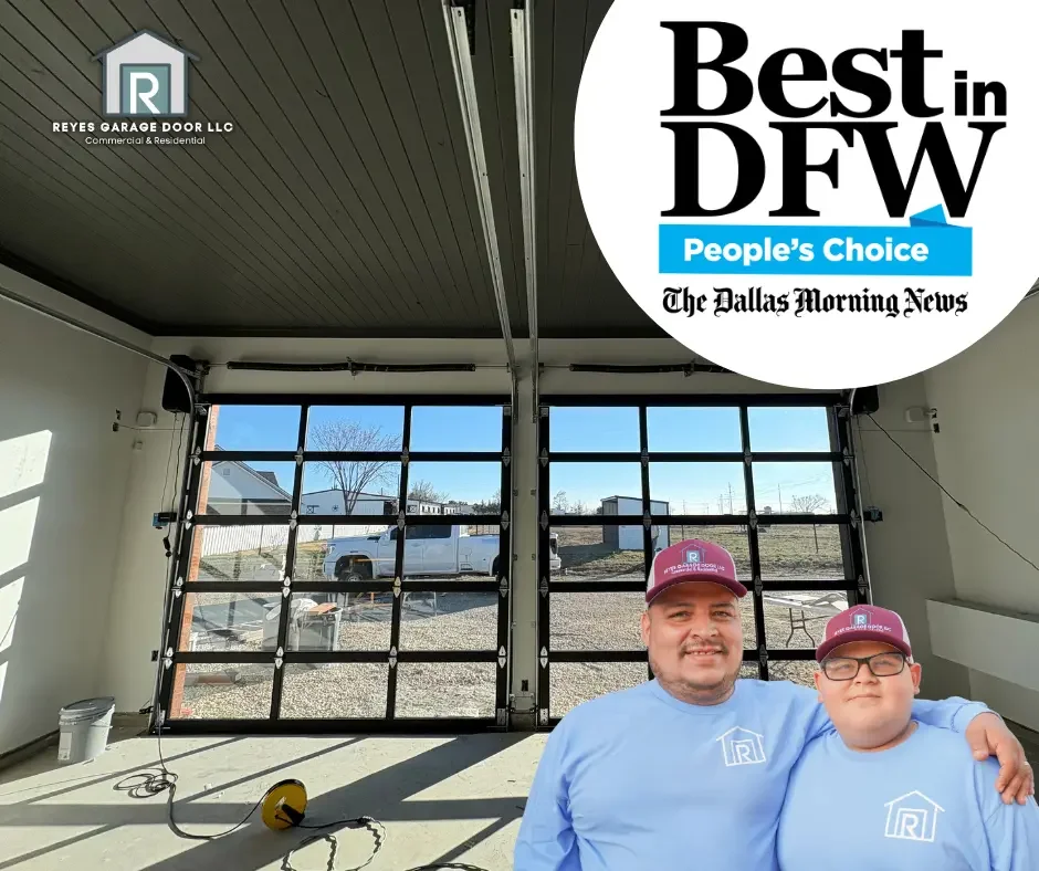 Two men wearing matching light blue shirts and maroon caps with a logo, standing inside a garage with glass front doors, outdoor scene visible through the doors, and a partially visible yellow electrical cord on the floor.