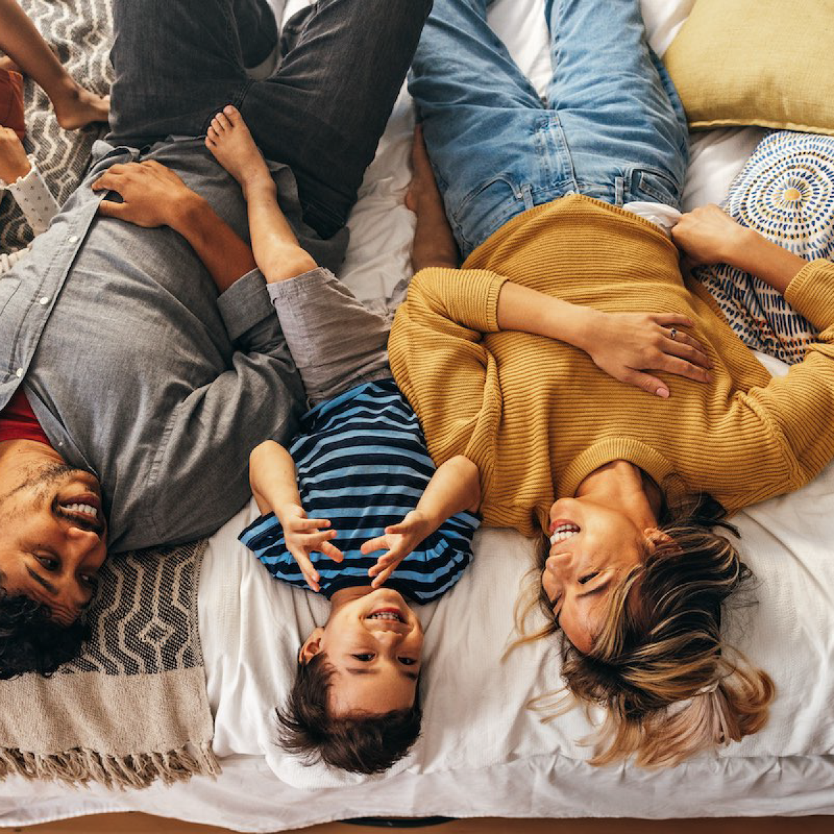 A family of three, including a woman, a man, and a young boy, lying on a bed and laughing together.