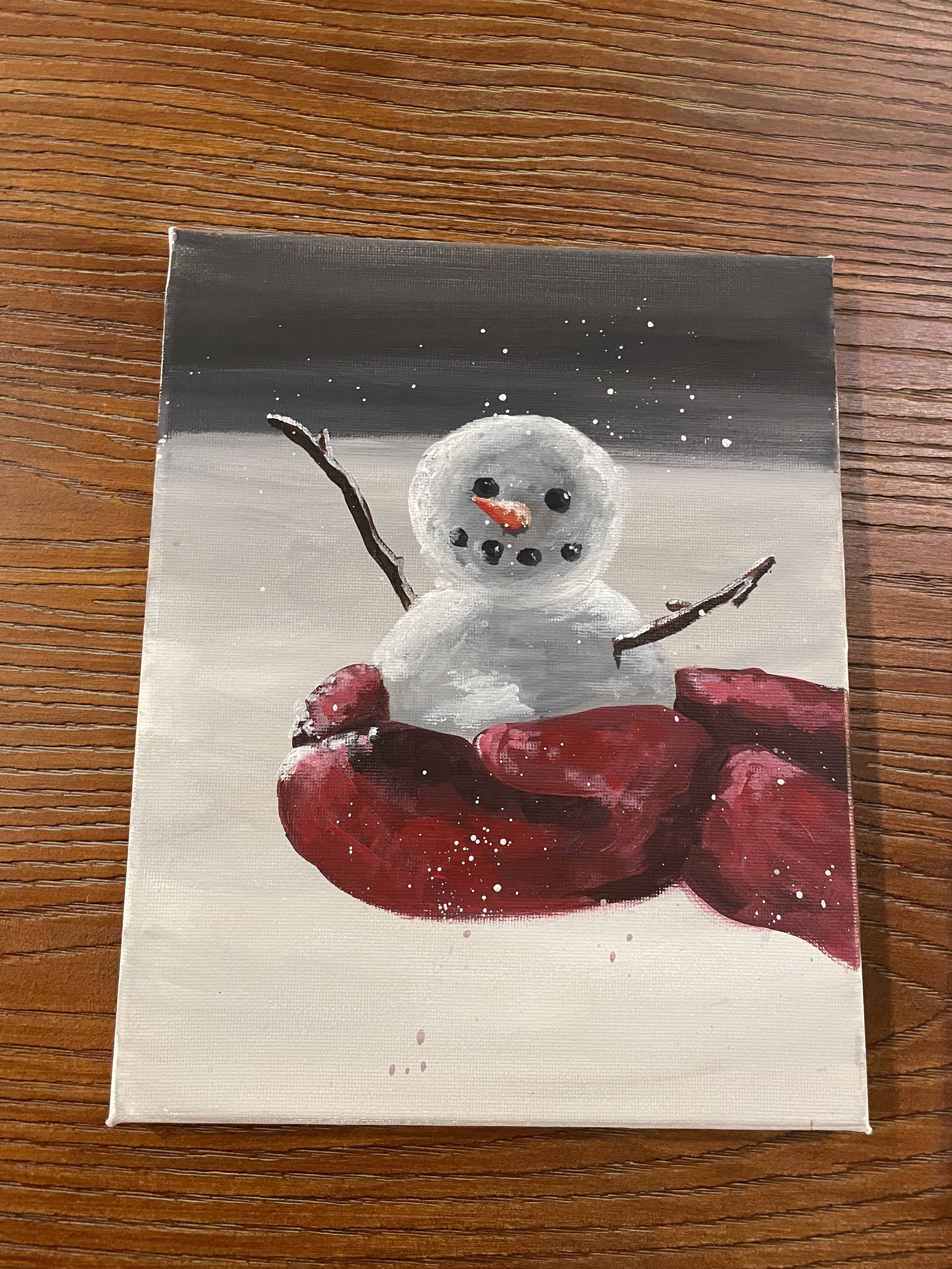 Painting of a snowman with a carrot nose, black eyes, and black buttons, standing on red rocks against a snowy background with snowflakes.