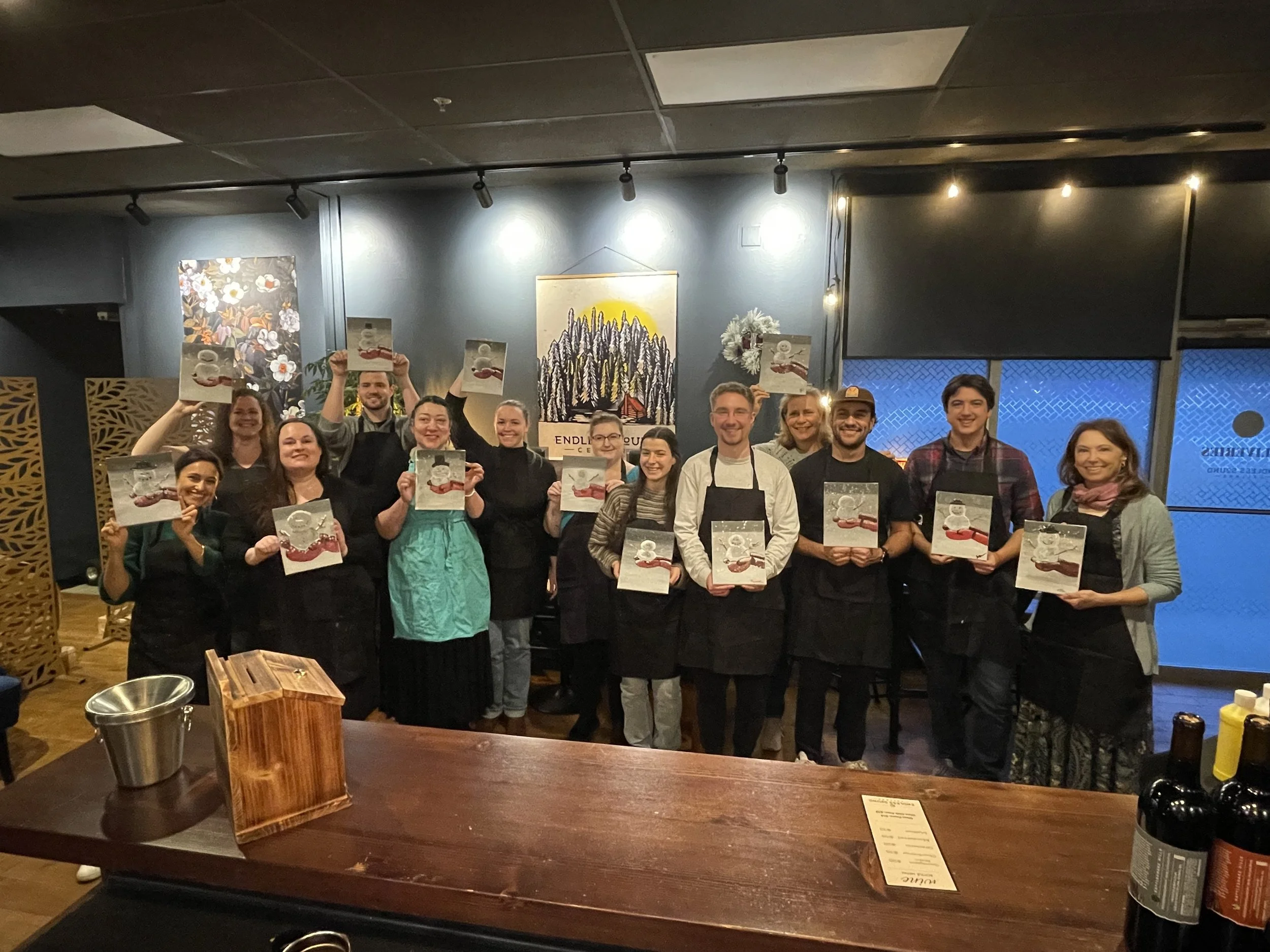 Group of people at a cooking class holding recipe books, smiling in a restaurant with artwork on the wall behind them. Paint and Sip class led by Amira Elsokkary in Olympia, WA