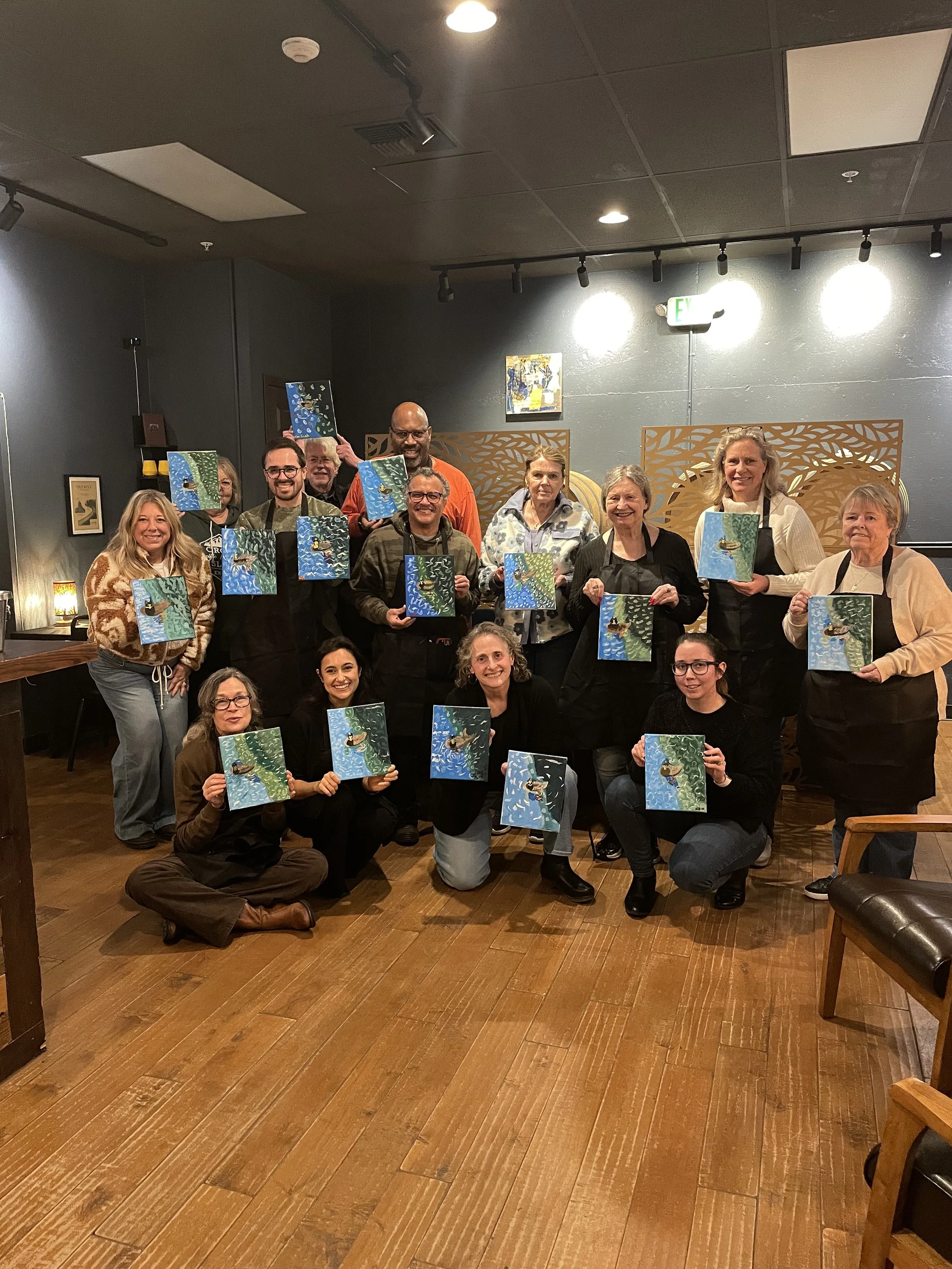 Group of people posing together in a restaurant, each holding a painting of a forest scene with a boat on a river.