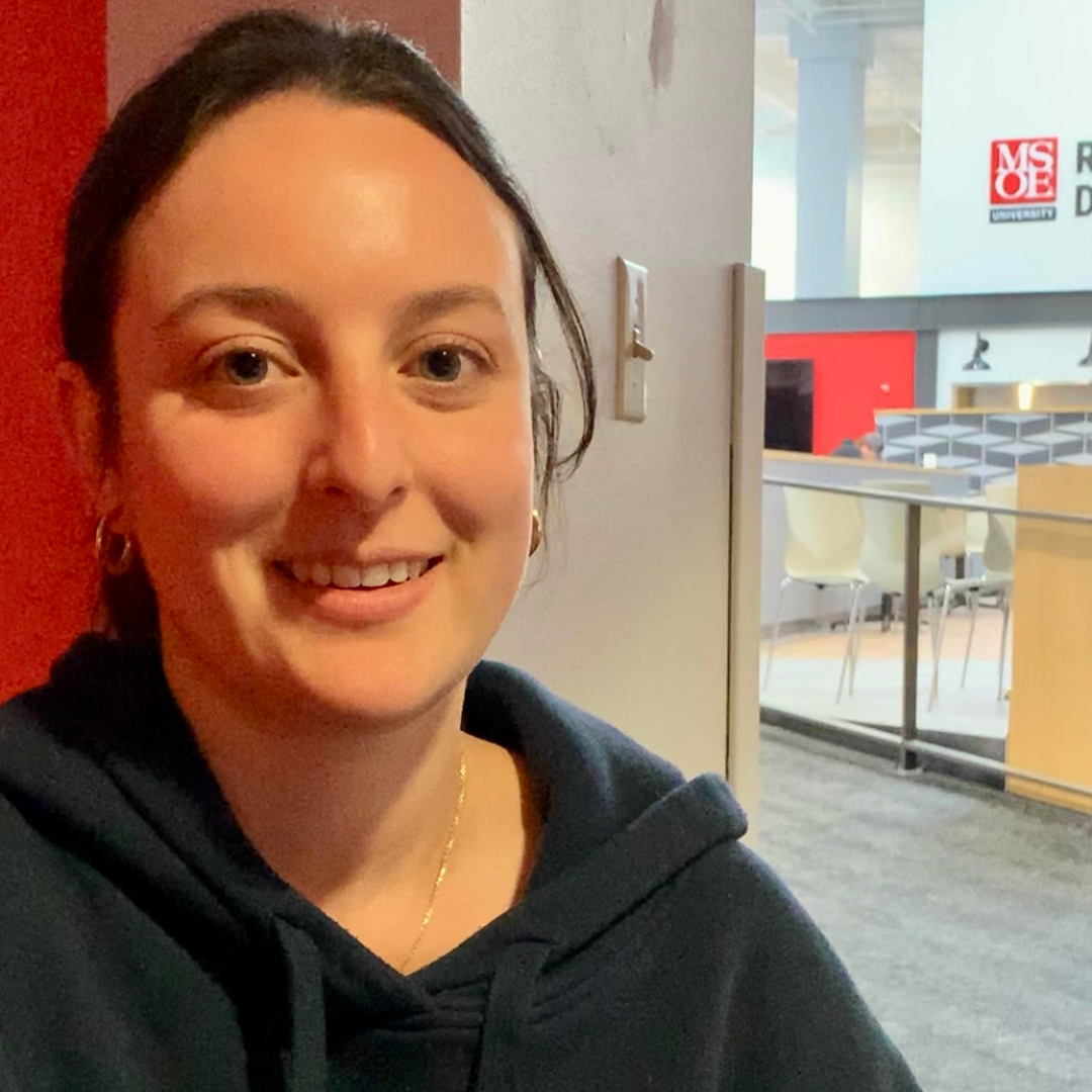 A person smiling indoors next to a red wall, with chairs and tables in the background.