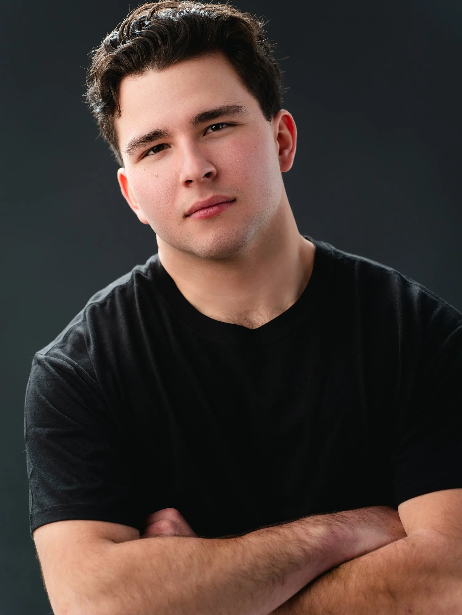 Close-up portrait of a young man with dark hair, wearing a black T-shirt, with arms crossed, against a dark background.
