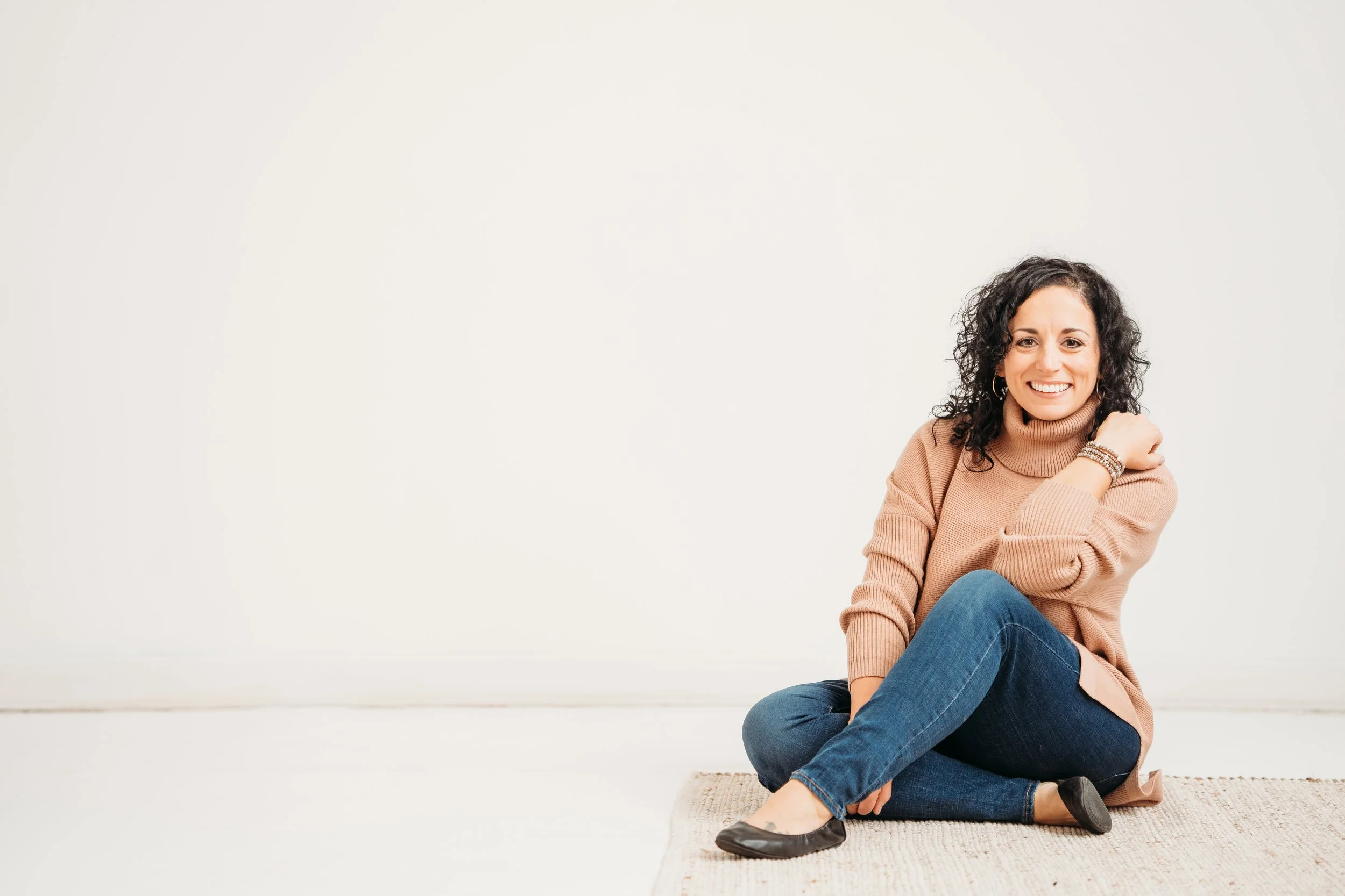 Woman sitting on a rug, wearing a tan sweater and jeans, smiling against a white background.