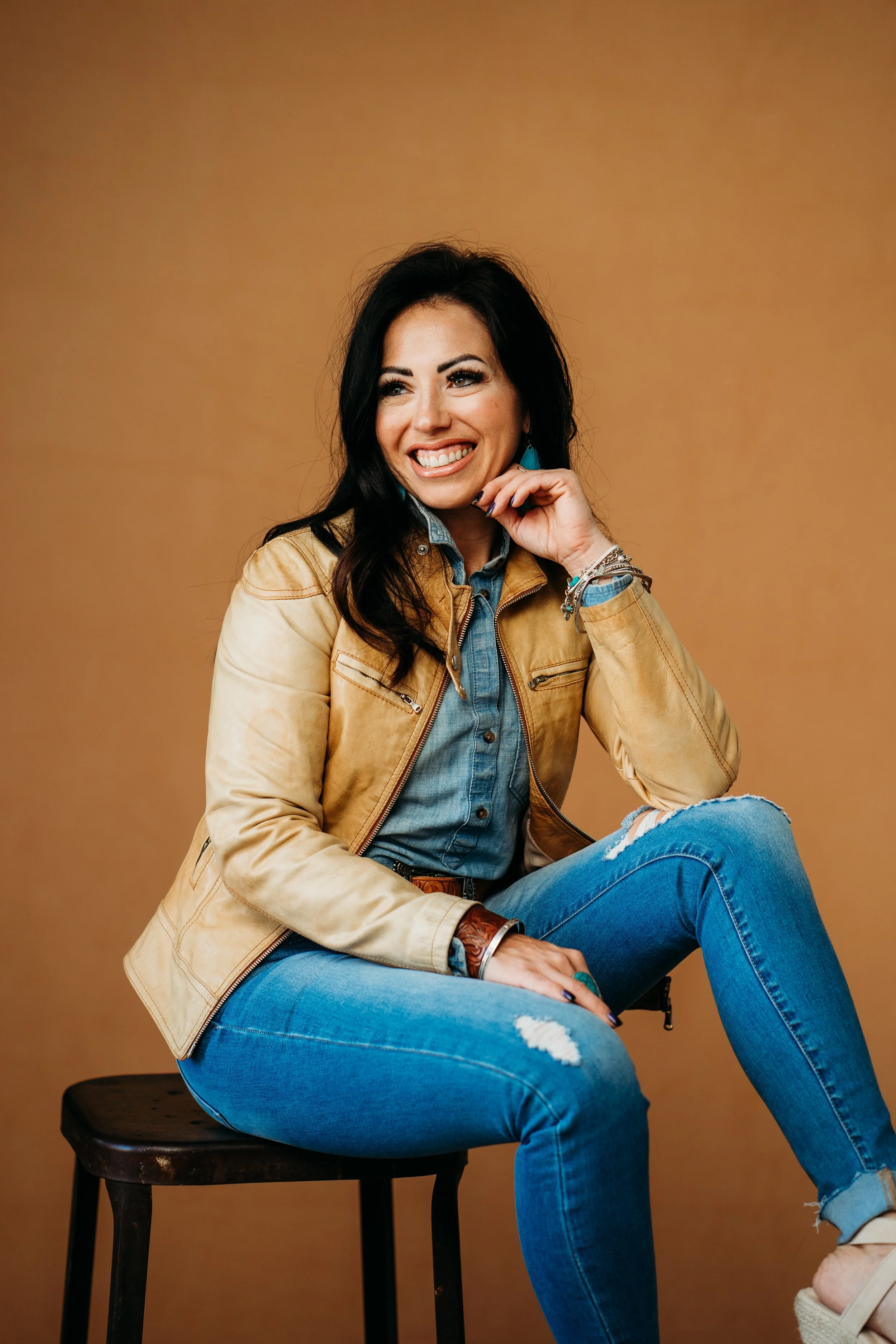 Smiling woman sitting on a stool, wearing a tan leather jacket over a denim shirt and blue jeans, with bracelets on her wrist, against a brown background.