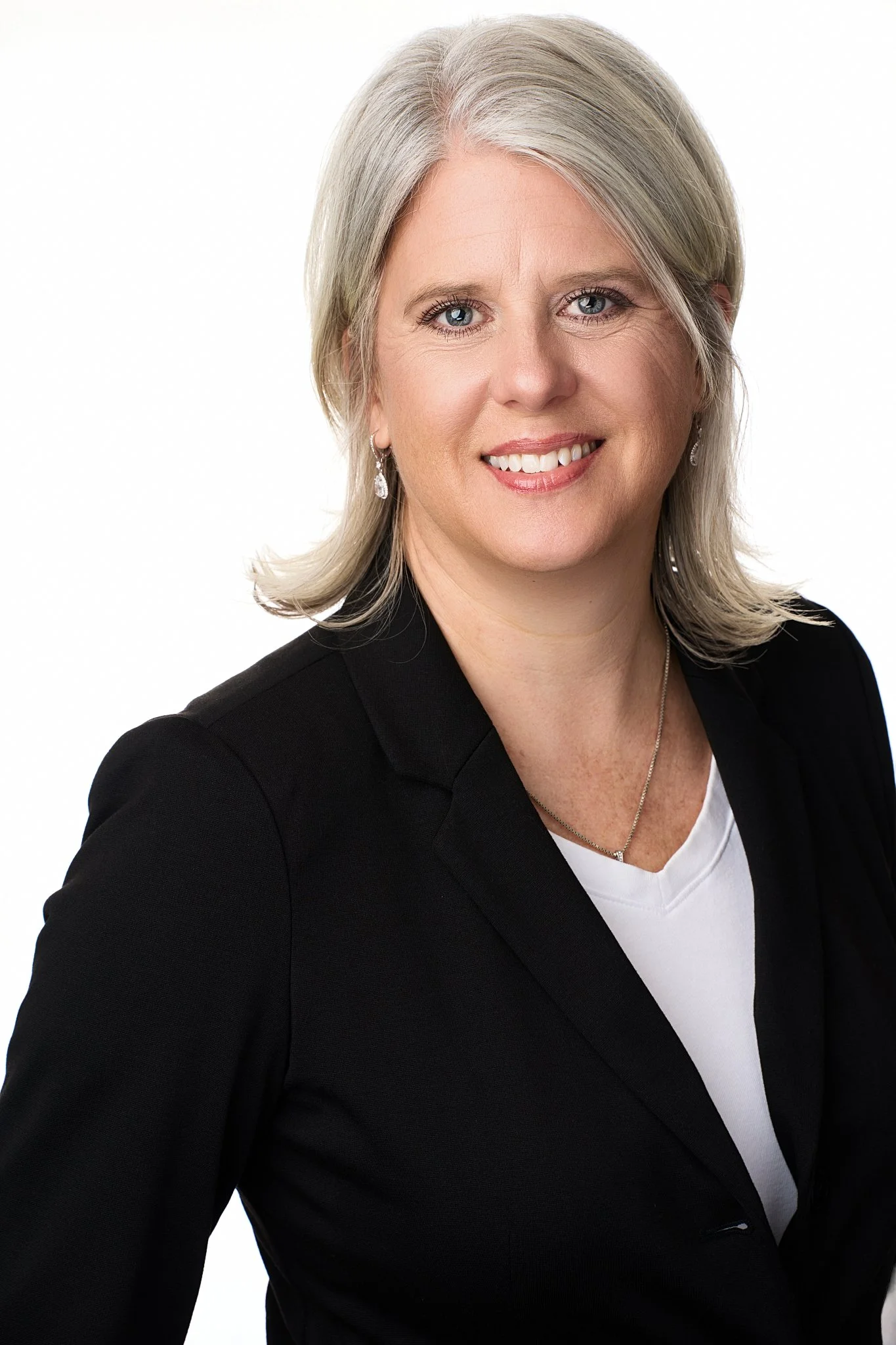 Smiling woman with shoulder-length grey-blonde hair wearing a black blazer over a white top, photographed against a light grey background.