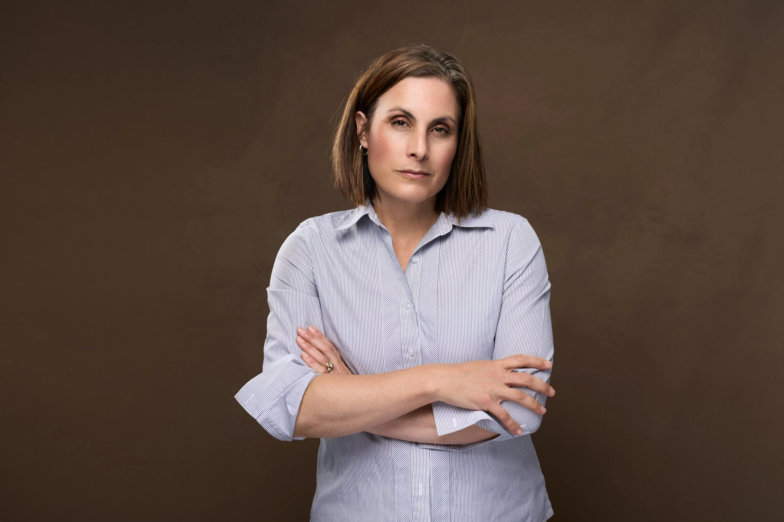 Woman with shoulder-length brown hair wearing a light blue and white pinstriped button-down shirt, arms crossed, against a warm brown backdrop.