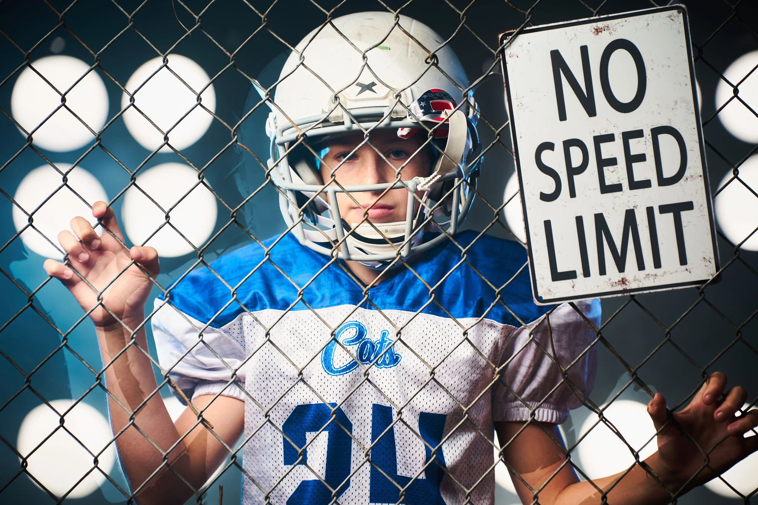 Youth football player in Cats jersey and helmet standing behind a chain-link fence, next to a 'No Speed Limit' sign, with stadium lights in the background