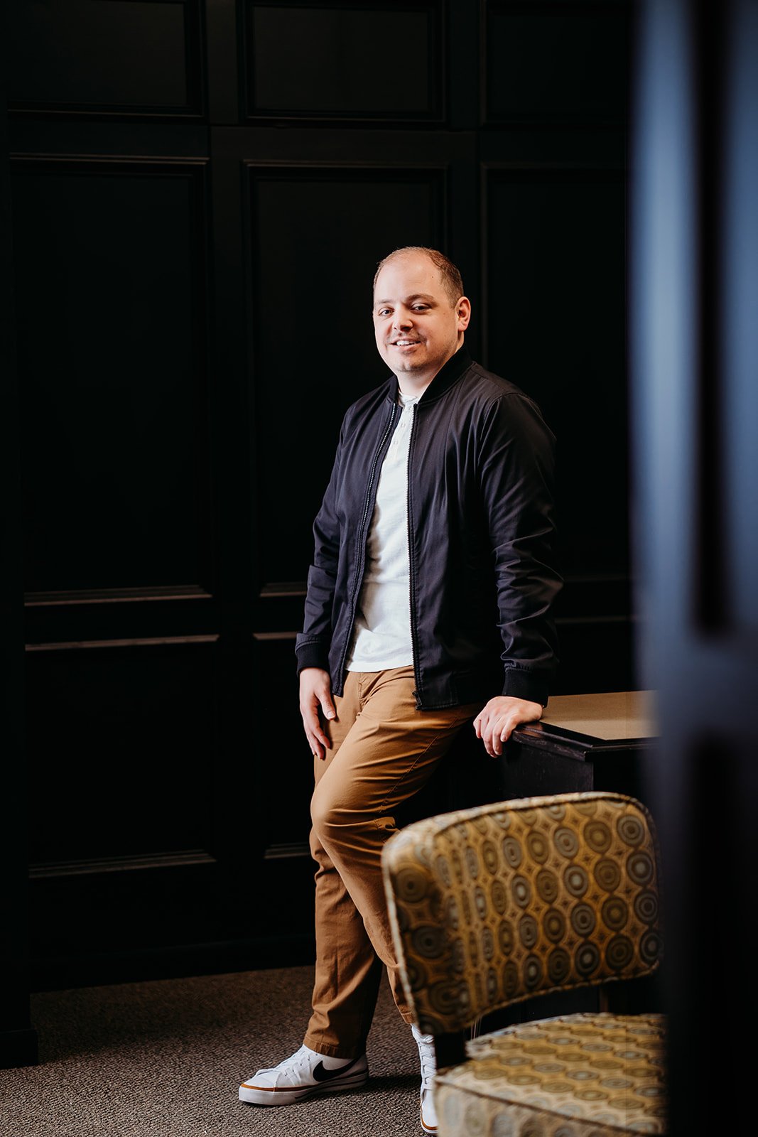 A man wearing a black jacket, white shirt, tan pants, and white Nike sneakers leaning against a small black table in a room with dark paneled walls.