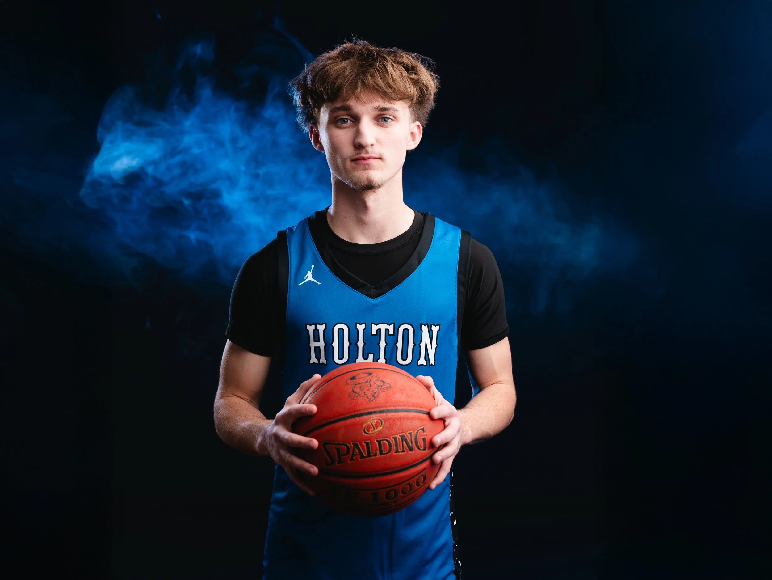 Young male basketball player holding a basketball, wearing a blue sports jersey with 'HOUTON' written on it, standing against a dark background with blue smoke.