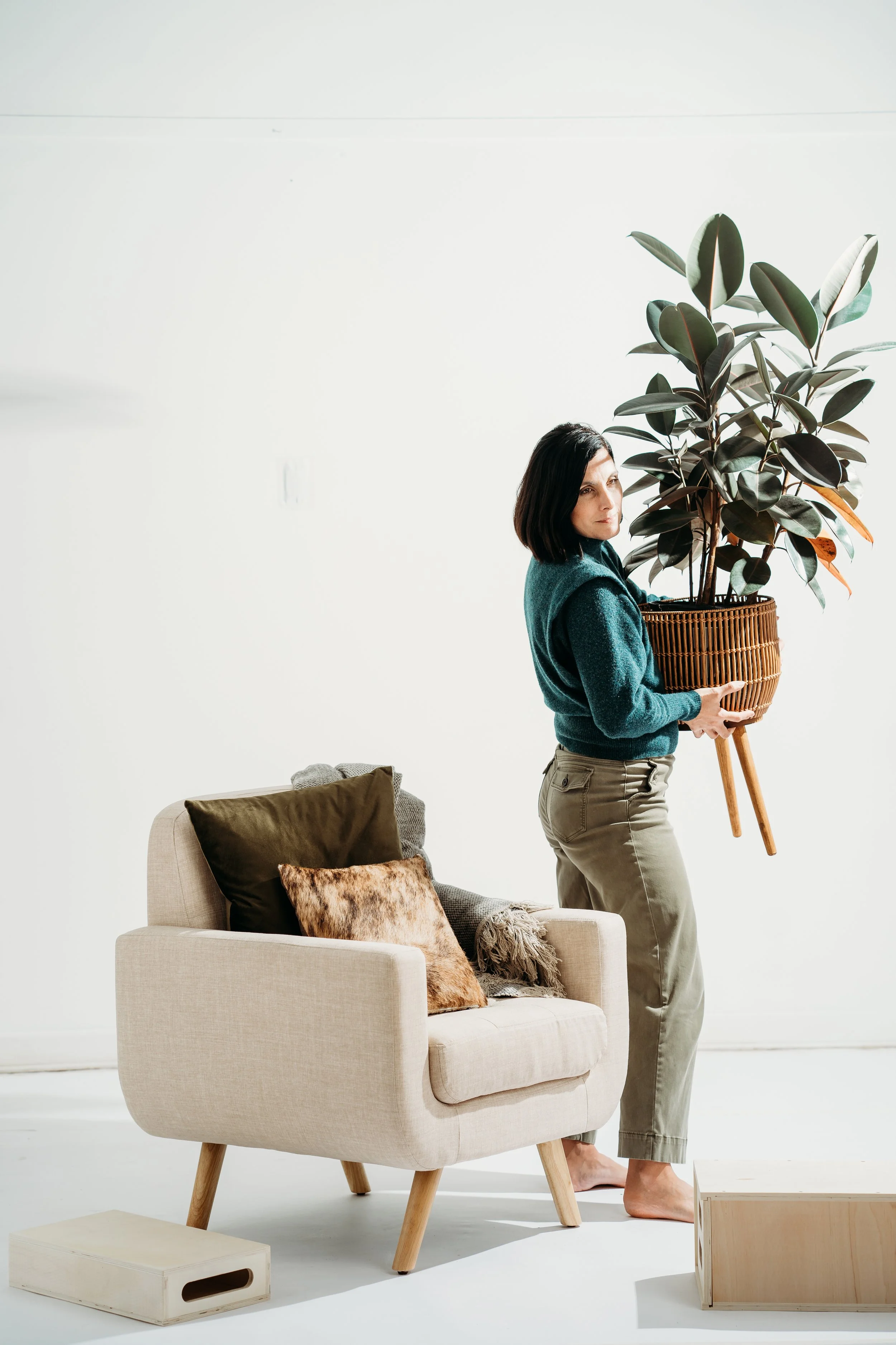 A woman holding a large potted plant with dark green leaves and wooden legs standing beside a beige sofa with three pillows, in a minimalist living room with a white wall.
