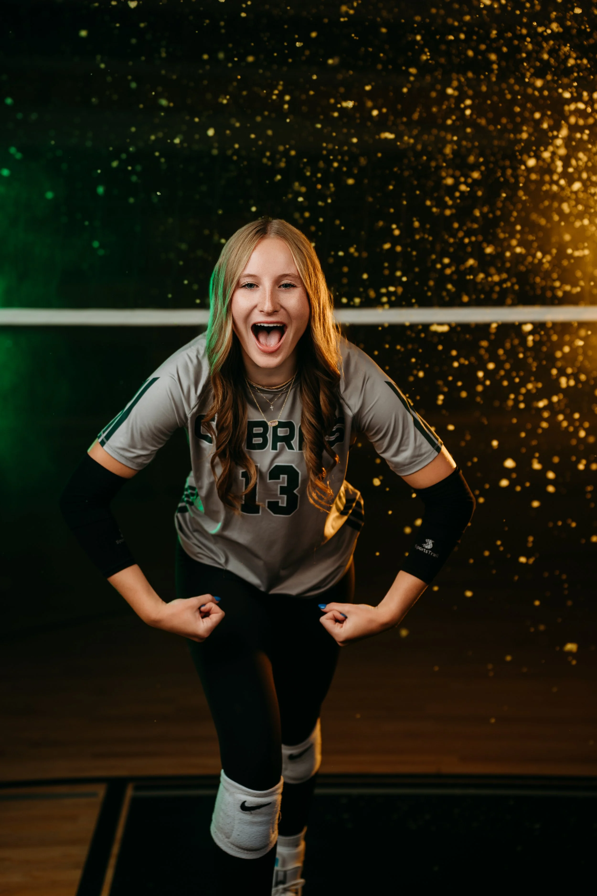 A female volleyball player in a gray jersey with the number 13, smiling and flexing her arms in a victory pose against a black background with gold and green confetti-like particles.