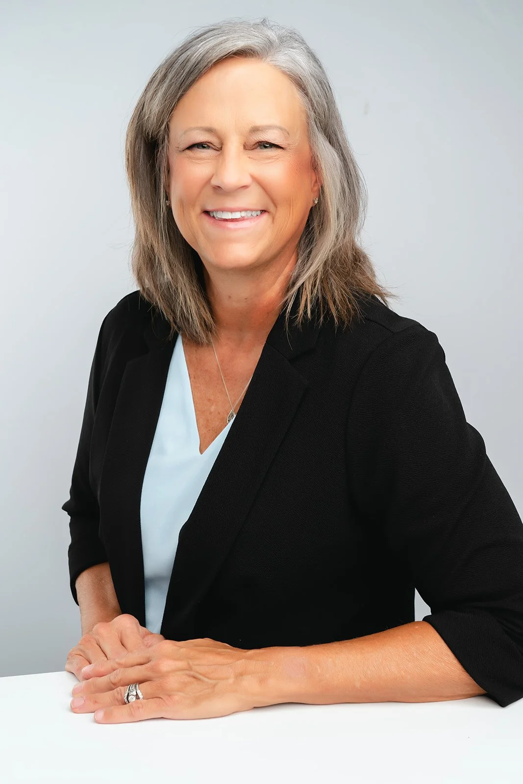 A smiling middle-aged woman with shoulder-length gray hair, wearing a black blazer over a light blue top, sitting at a white desk with her hands resting on it, in a professional portrait studio setting.