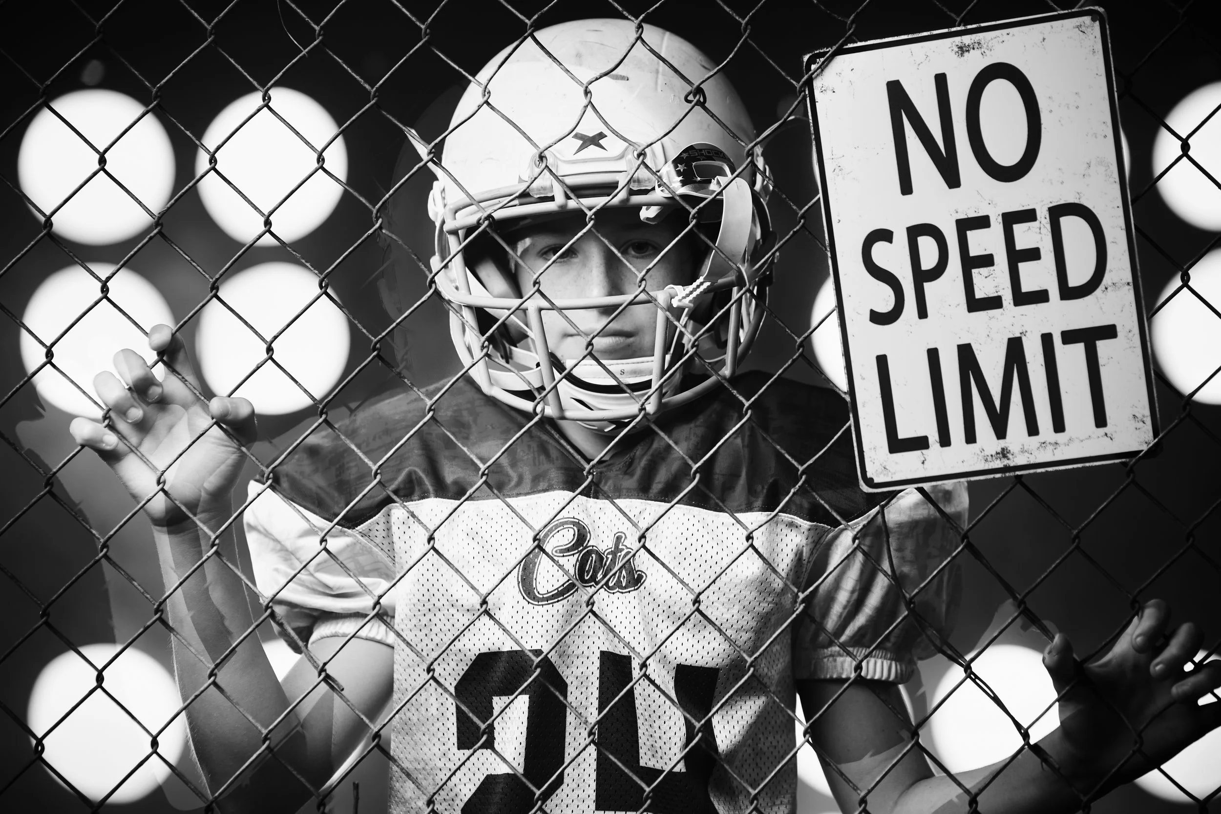 Youth football player in Cats jersey and helmet standing behind a chain-link fence, next to a 'No Speed Limit' sign, with stadium lights in the background in black and white