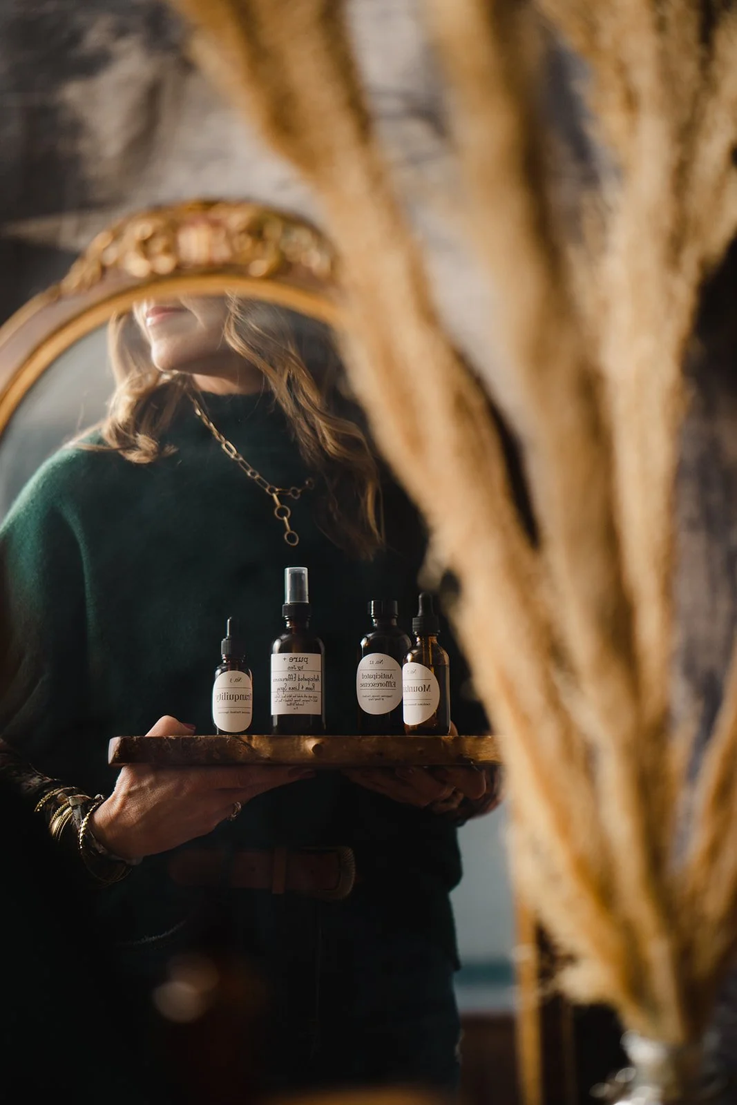Woman holding a tray with skincare bottles and spray behind a mirror